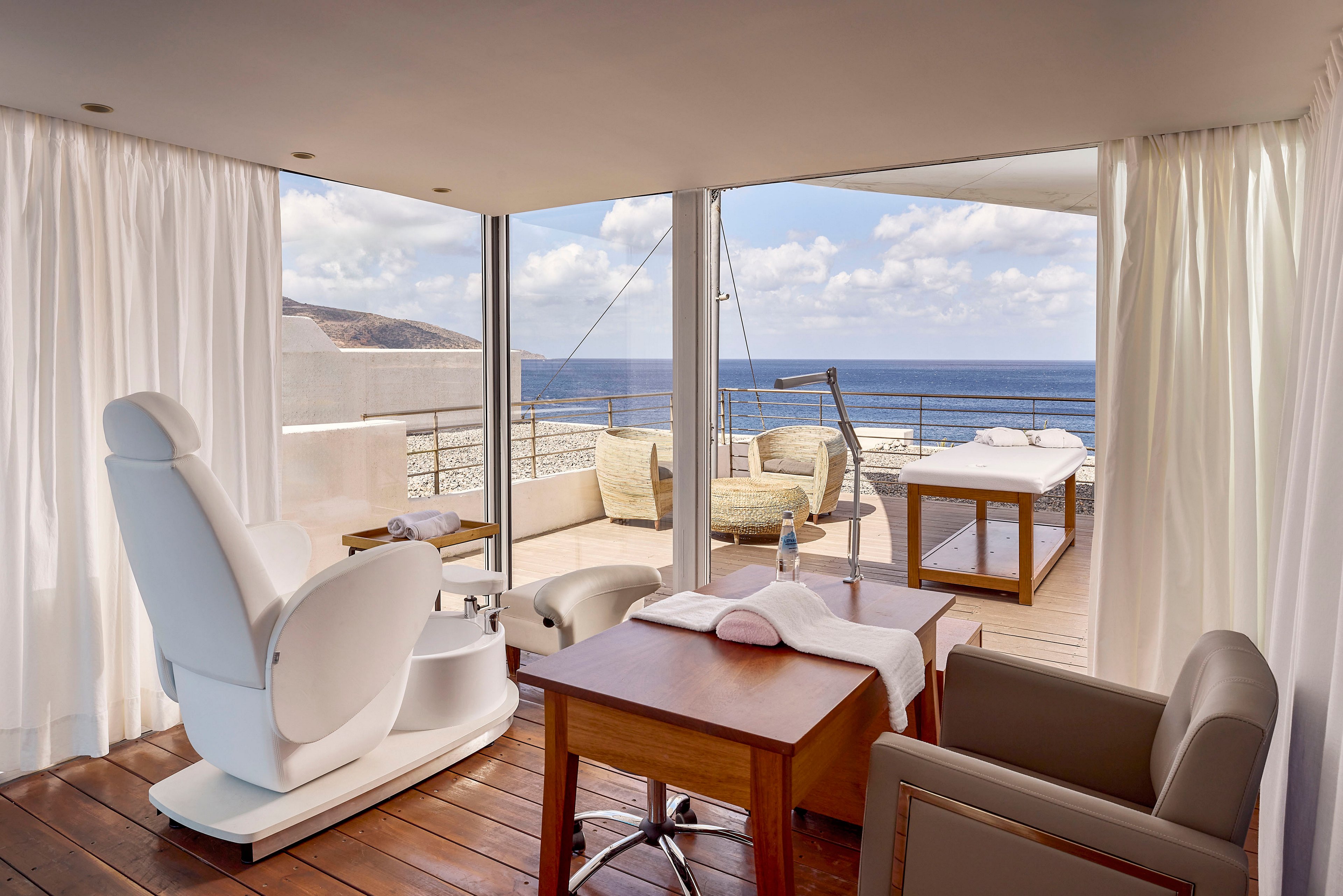 A modern spa treatment room features a reclining white chair and a wooden desk set on a polished wooden floor. Large glass doors open onto a terrace with lounge chairs and a massage table overlooking the ocean and distant coastline. Soft white curtains frame the space, creating a serene and private atmosphere. The bright natural light and neutral tones emphasize relaxation and coastal luxury.