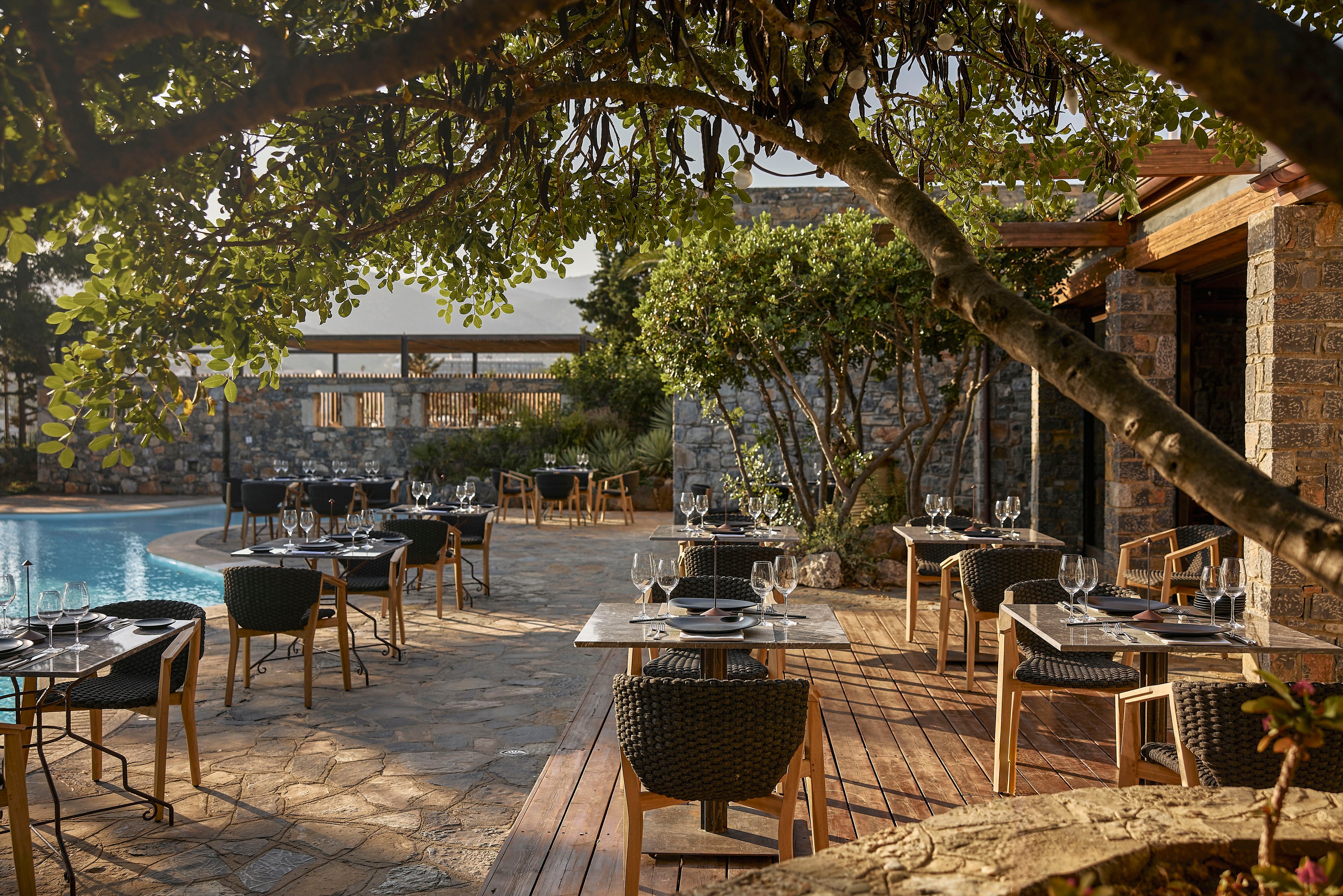 An elegant outdoor restaurant terrace is set with tables and chairs beside a curved swimming pool. Each table is neatly arranged with glassware and cutlery, ready for guests. Stone walls and wooden structures frame the space, while lush trees and plants provide shade and greenery. Warm sunlight creates a relaxed, upscale atmosphere ideal for al fresco dining.
