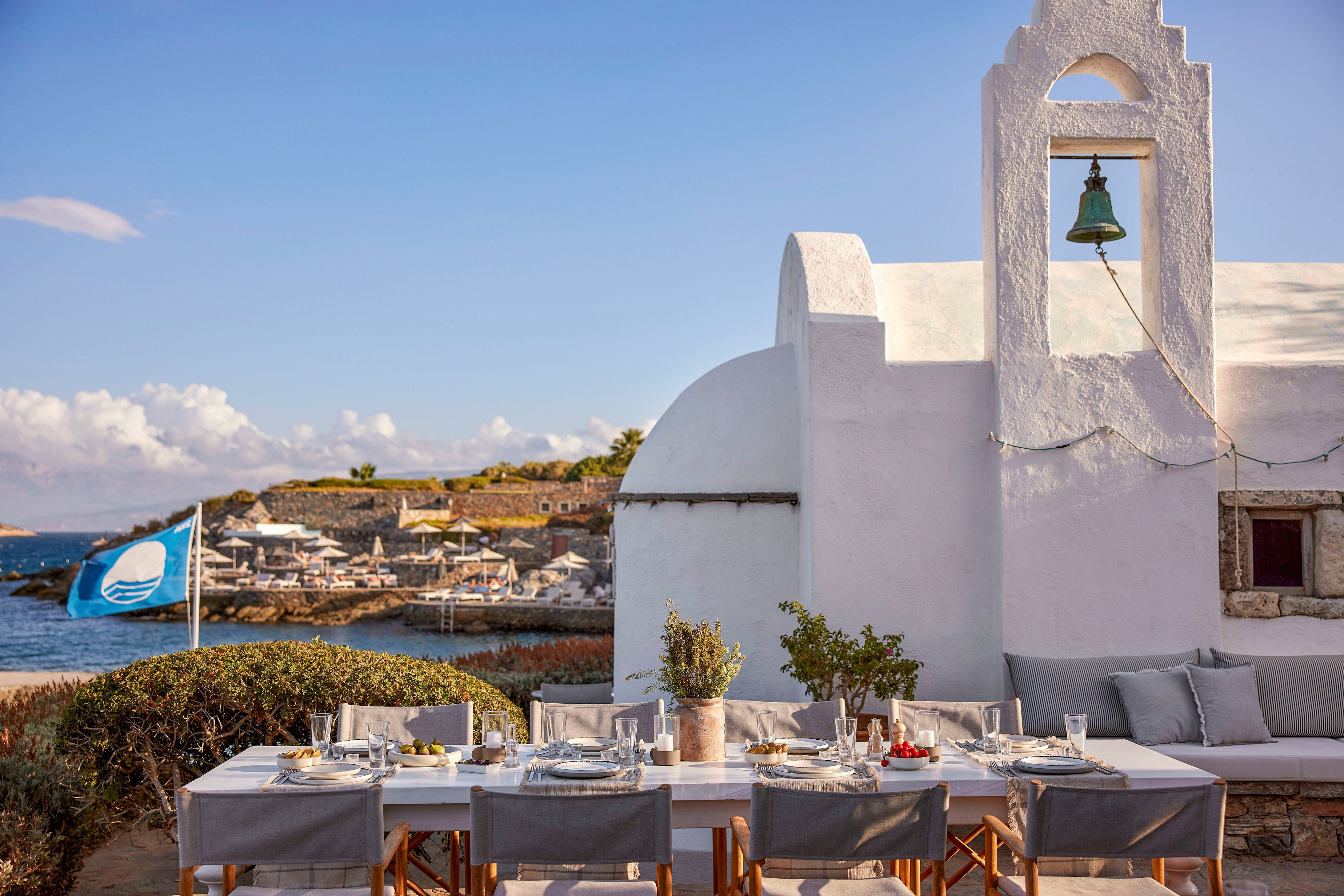 An elegant outdoor dining table is set beside a whitewashed chapel overlooking the sea. The table is arranged with plates, glasses, cutlery, and neutral-toned textiles, ready for guests. In the background, a coastal resort with umbrellas and loungers lines the shoreline under a clear sky. A blue flag waves near the water, and a bell hangs in the chapel tower, adding to the Mediterranean atmosphere.