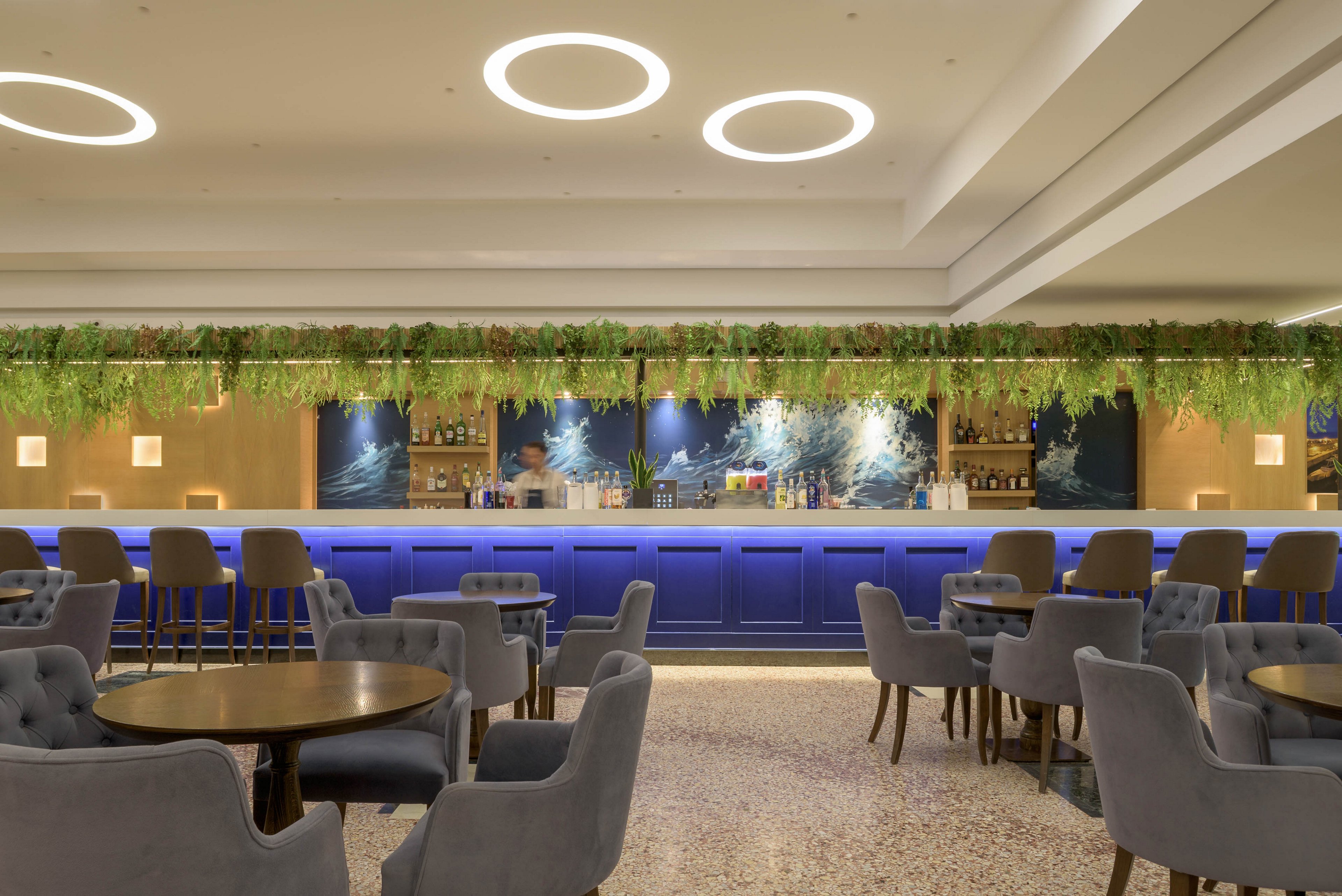 A contemporary hotel bar features a long blue counter with a variety of bottles and glassware displayed behind it. Grey upholstered chairs and round wooden tables fill the foreground, creating a comfortable lounge area. Circular ceiling lights and warm wall sconces provide soft, even illumination. A hanging strip of greenery runs above the bar, adding a natural accent to the sleek interior design.