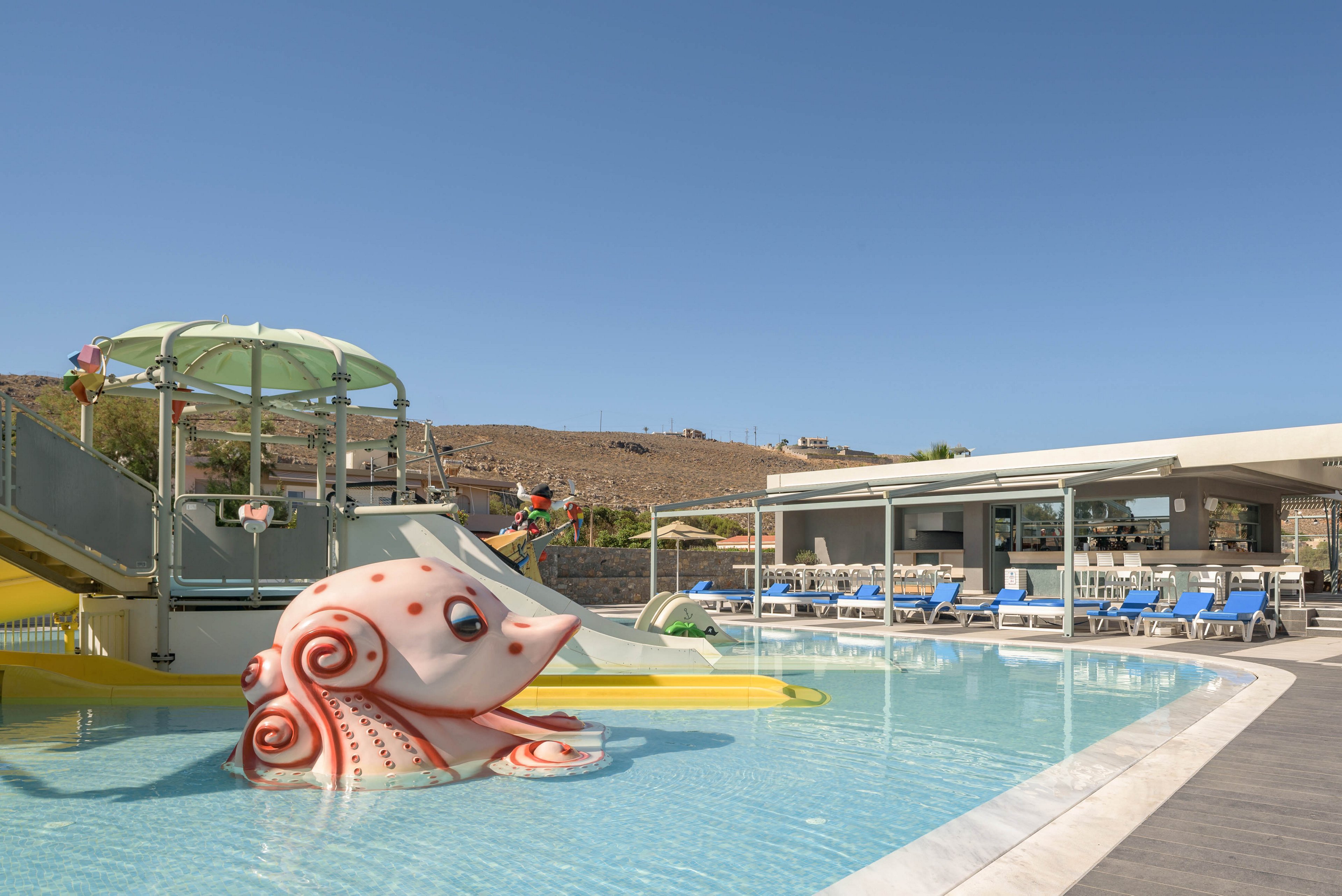 An outdoor resort swimming pool features a playful octopus sculpture in shallow water. A small water slide structure with a canopy and stairs stands beside the pool, designed for children. Blue lounge chairs line the poolside terrace near a modern building with large windows. Dry hills and a clear blue sky create a sunny vacation atmosphere in the background.