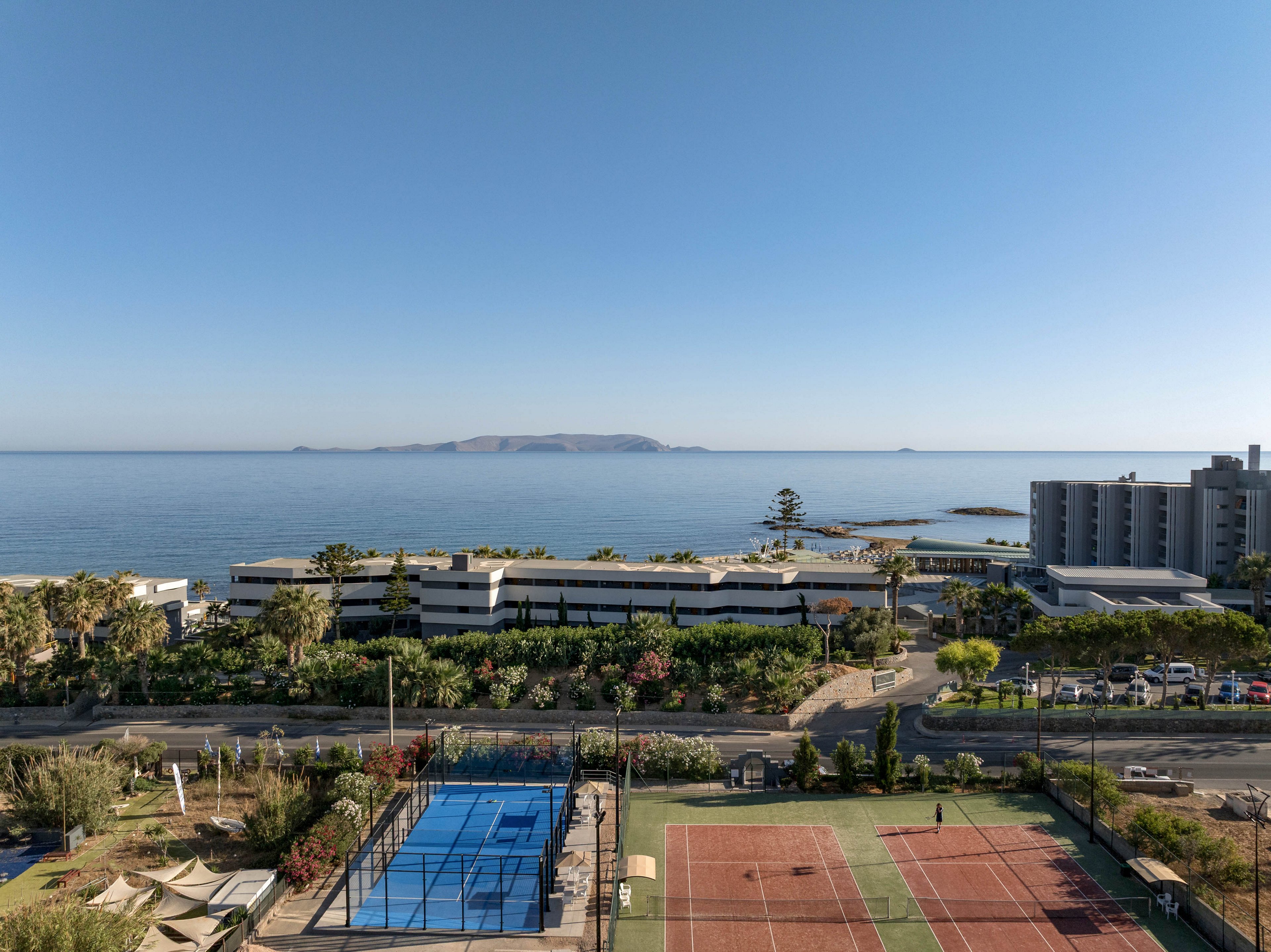 A seaside resort complex overlooks a calm blue ocean with an island visible on the horizon. In the foreground, outdoor tennis and padel courts are surrounded by greenery and palm trees. A road separates the sports area from low-rise hotel buildings and landscaped gardens. The clear sky and bright daylight create a relaxed vacation atmosphere along the coastline.