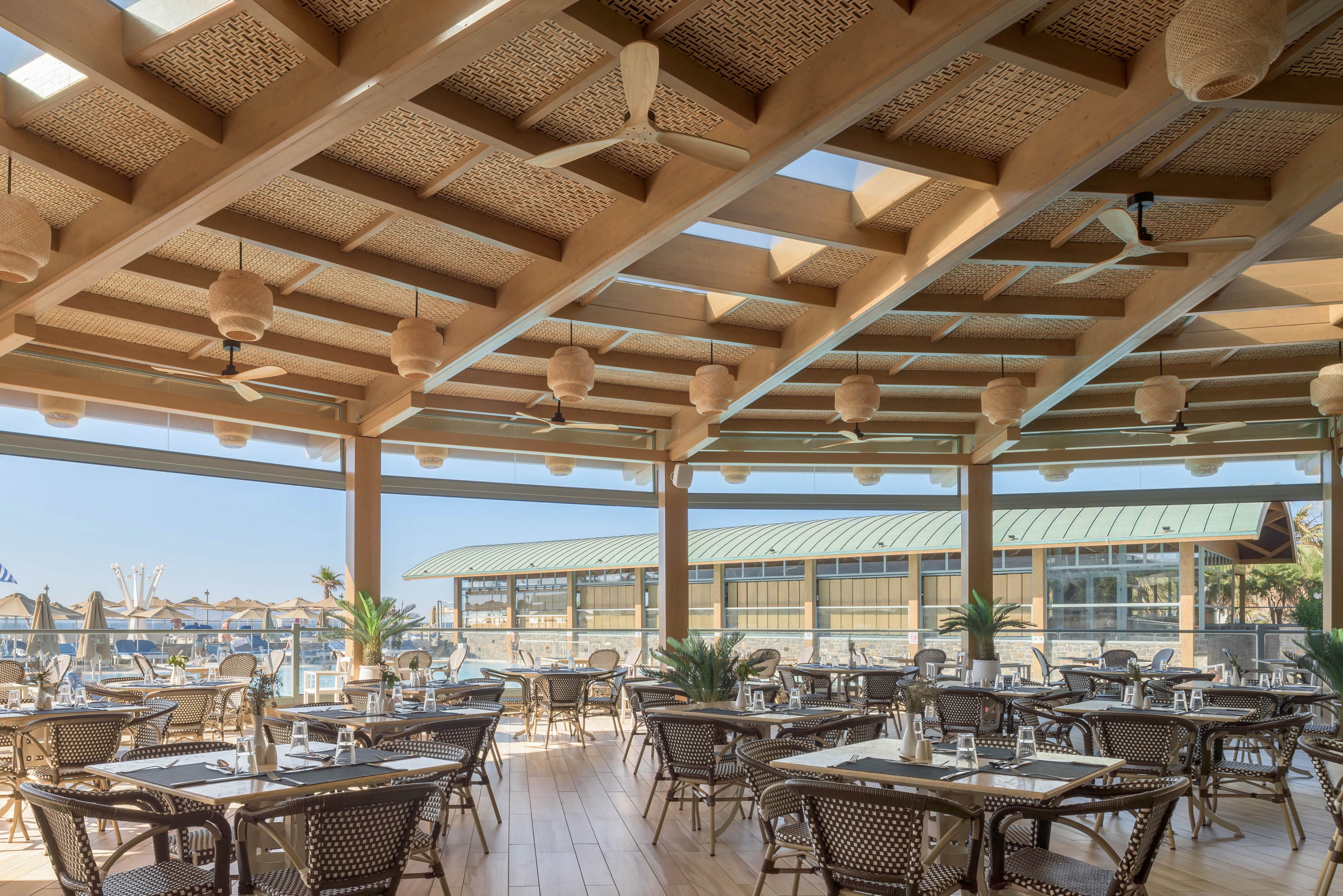 An open-air restaurant features neatly arranged tables and chairs beneath a large wooden pergola. The space overlooks a waterfront area with boats and a long building with a green metal roof in the background. Natural light floods the dining area, highlighting the warm wood flooring and woven ceiling details. Hanging lanterns, ceiling fans, and potted plants contribute to a relaxed, coastal atmosphere.