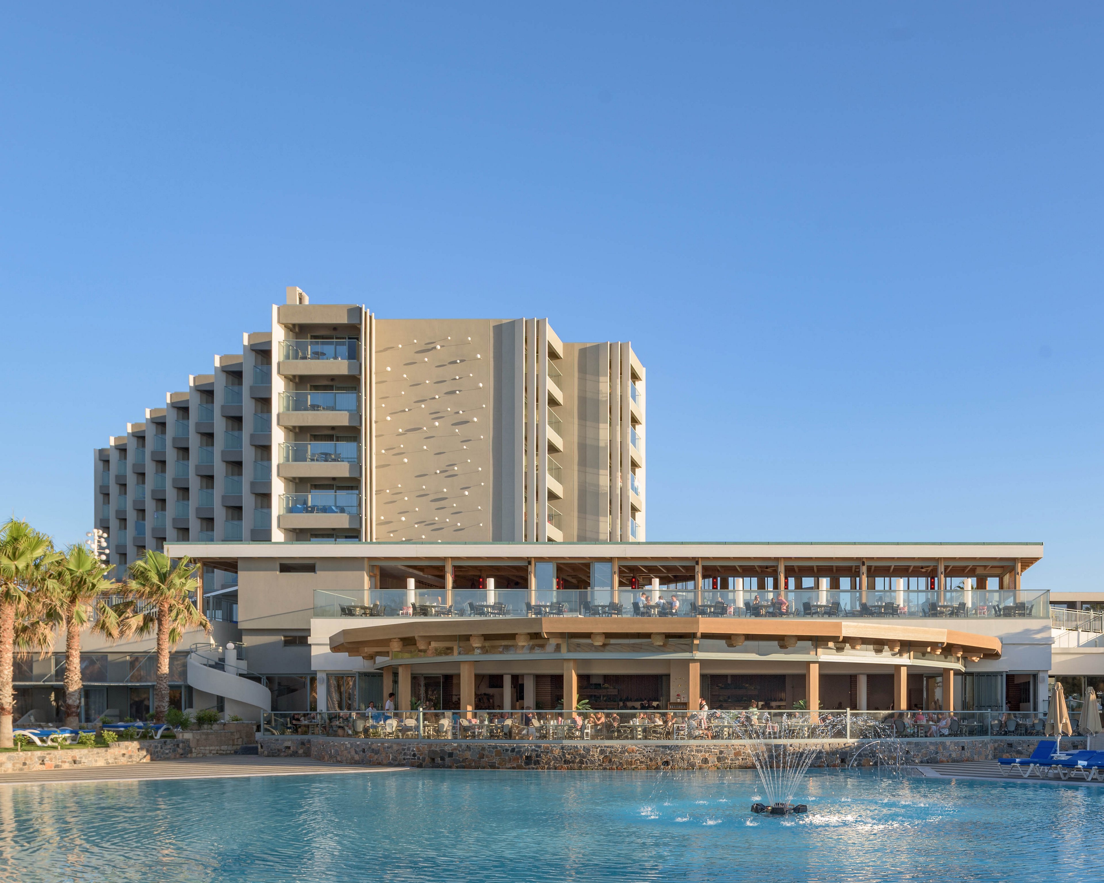 A large modern resort hotel stands beside a calm, blue swimming pool. The multi-story building features balconies and a central vertical facade detail, with a lower circular structure in the foreground. Guests can be seen relaxing near the water, while a small fountain sprays in the pool. Palm trees and clear blue sky emphasize the warm, coastal setting.