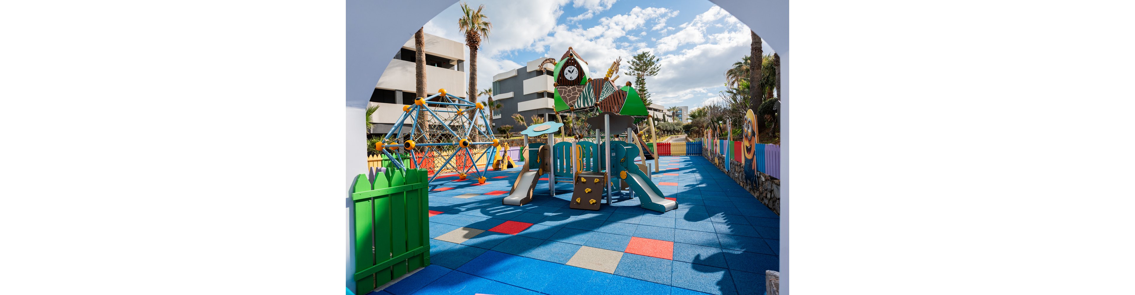 The image shows a vibrant outdoor playground featuring a slide, climbing frame, and geometric climbing dome. The setting is surrounded by colorful fencing and soft blue flooring tiles with red and beige accents, located near modern buildings and palm trees. The playground is designed for children, with playful visuals and a cheerful atmosphere under a partly cloudy sky.