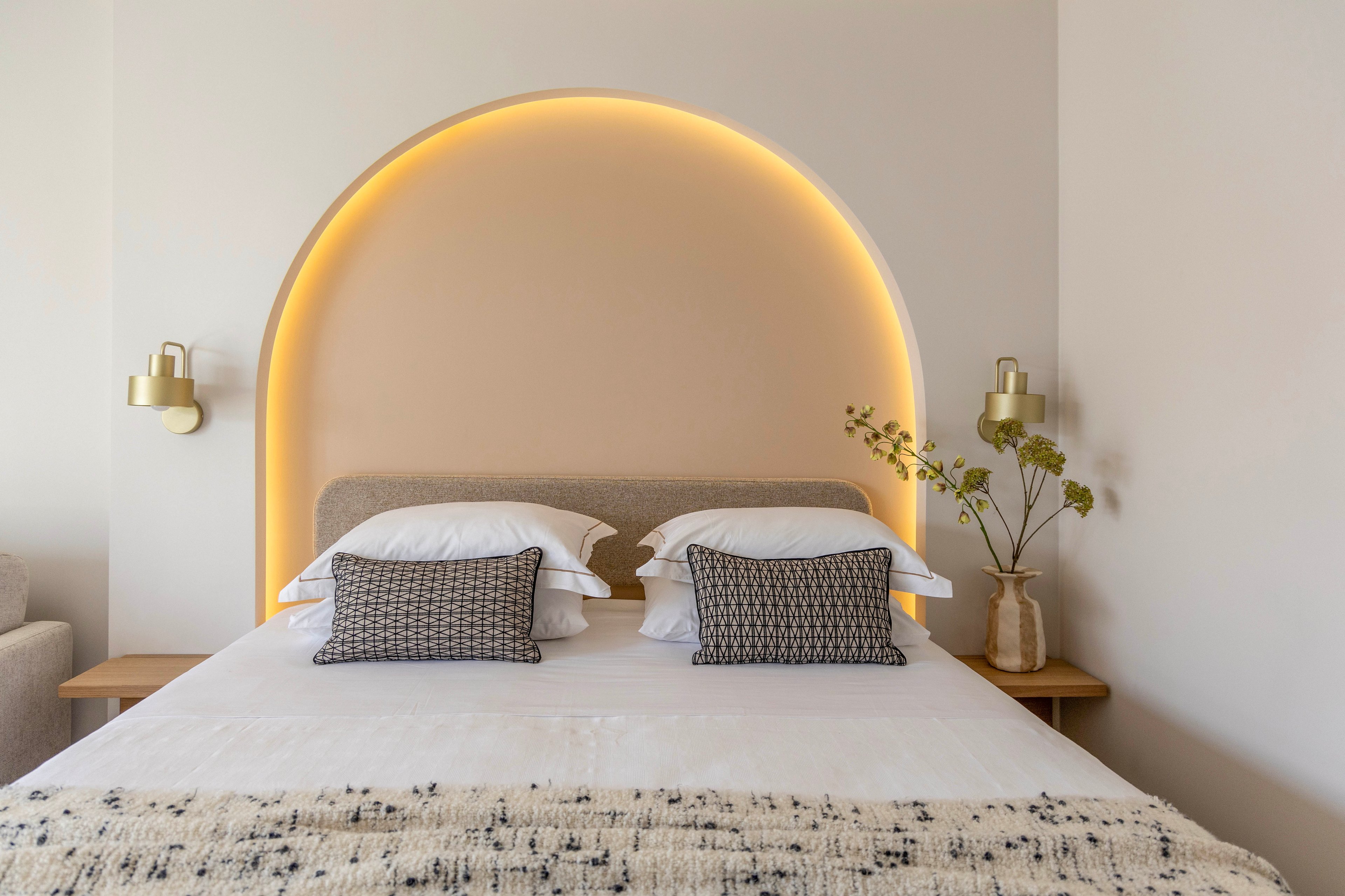 A neatly made bed with layered white pillows and patterned cushions sits centered beneath a softly glowing arched wall light. Minimalist sconces flank the bed, adding to the contemporary design of the room. A textured throw blanket and neutral-toned headboard contribute to the cozy, inviting atmosphere. A small bedside table with a ceramic vase and greenery completes the calm, modern interior setting.