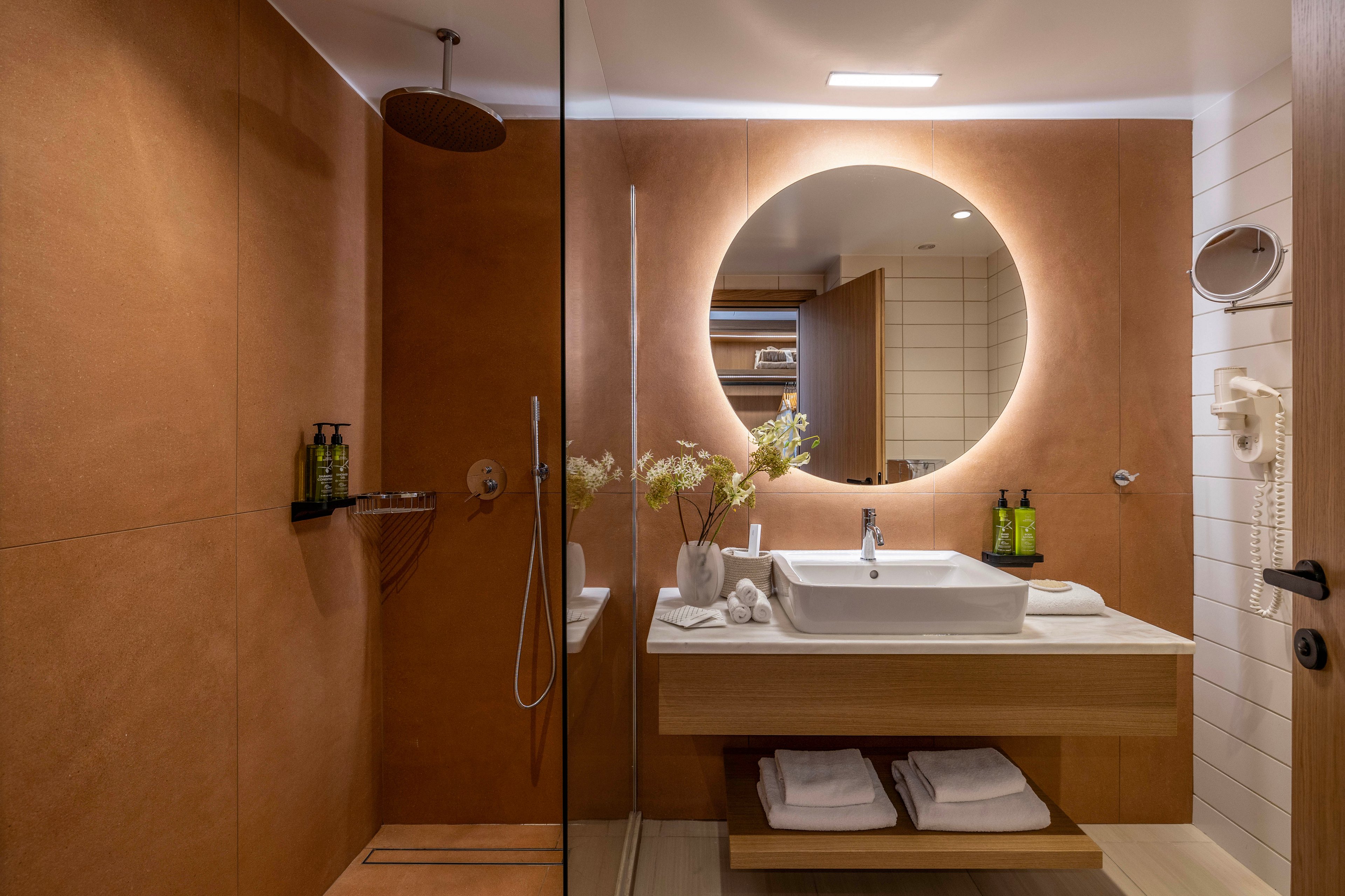 Contemporary bathroom interior featuring a large round backlit mirror above a rectangular countertop sink. A walk-in shower with a rainfall showerhead and glass partition is visible on the left, with toiletries arranged neatly. Warm brown wall panels contrast with white tiles and towels stored on the wooden vanity shelf. The space is softly lit, creating a cozy, upscale hotel atmosphere.