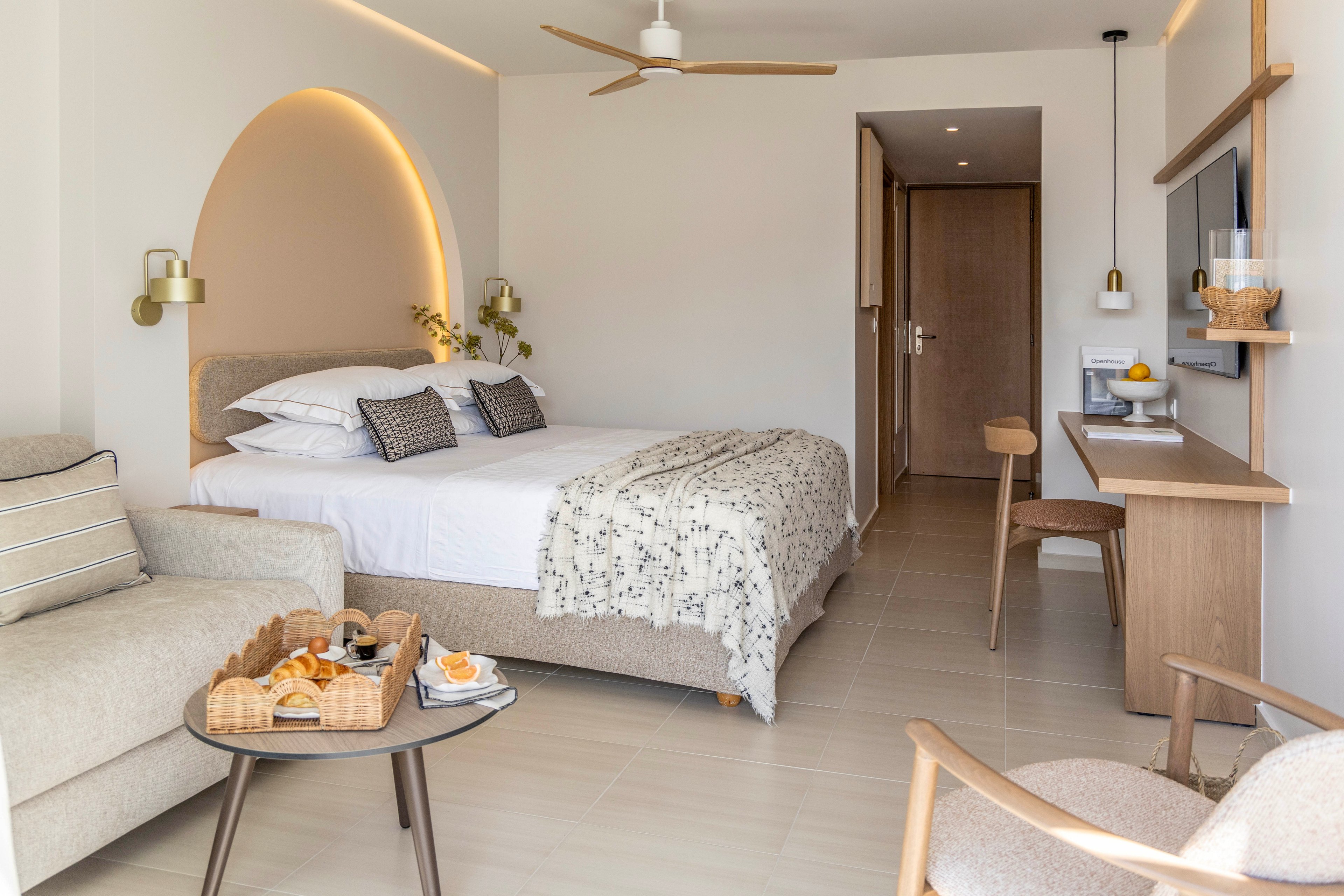 Contemporary hotel room interior featuring a neatly made double bed with patterned throw and accent pillows. A cozy sofa and armchair sit beside a small round table holding a breakfast tray with pastries, juice, and coffee. Light wood furniture, including a long desk and wall-mounted TV, complements the neutral color palette. Soft indirect lighting, minimalist decor, and a ceiling fan create an inviting, relaxing atmosphere.
