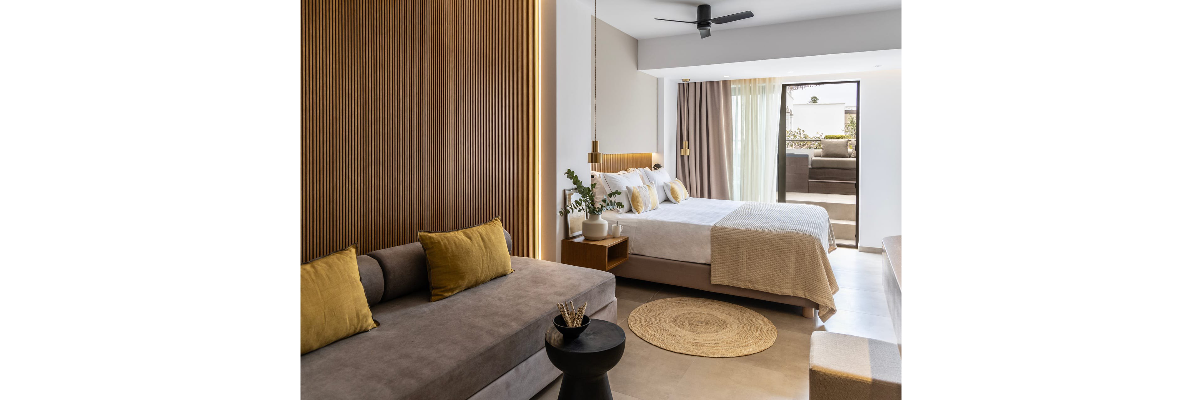 A contemporary hotel-style bedroom features a neatly made bed with neutral bedding and multiple pillows. In the foreground, a gray sofa with mustard-yellow cushions sits beside a small round side table. Vertical wooden wall paneling and warm accent lighting add texture and depth to the space. Large glass doors open to a sunlit terrace with additional seating, creating an airy and inviting atmosphere.