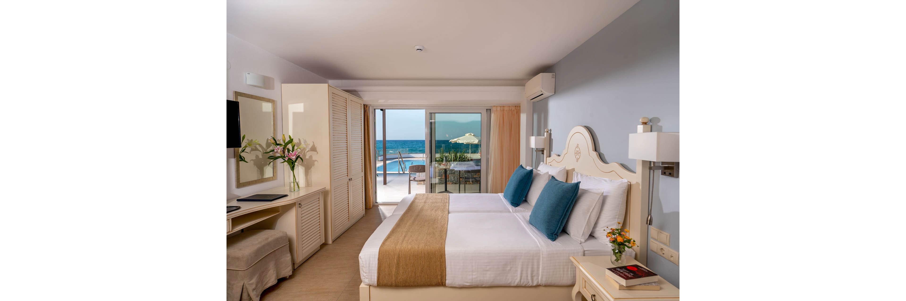 A bright hotel bedroom features a neatly made double bed with white linens and blue accent pillows. Large glass doors open onto a balcony overlooking the ocean, with outdoor seating and a sun umbrella visible. The room includes a wall-mounted television, a desk with a mirror, and a wardrobe on the left side. Fresh flowers and decorative details create a calm, inviting atmosphere ideal for a seaside vacation.