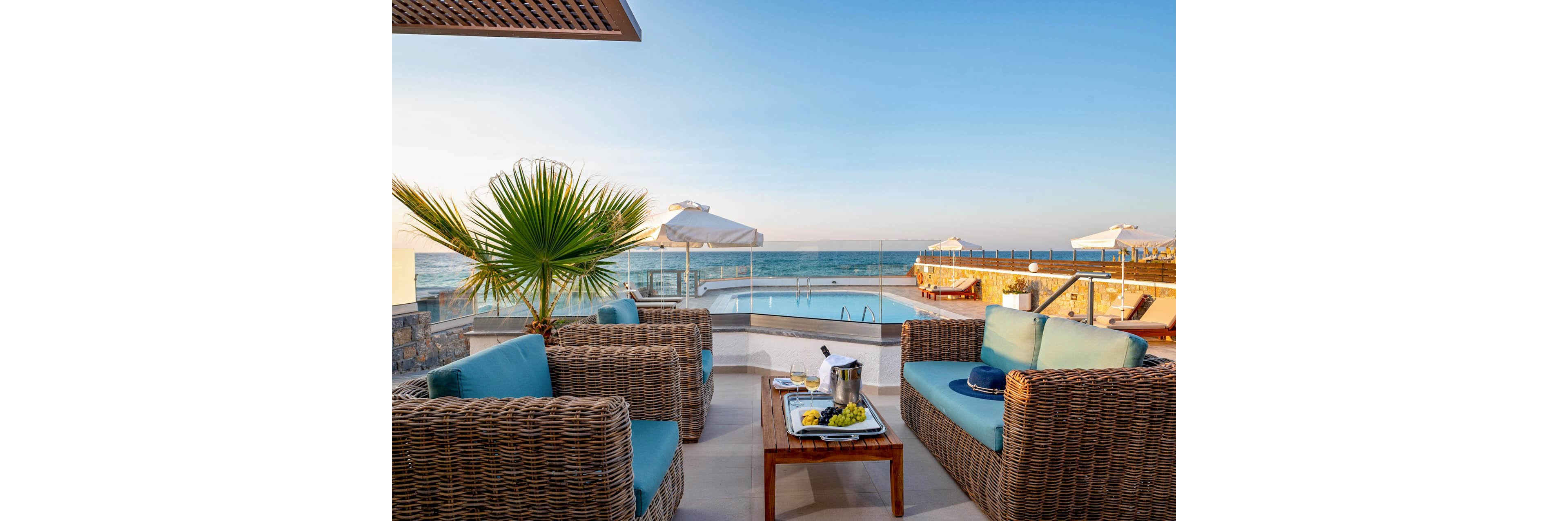 An inviting wicker lounge seating area overlooks a swimming pool and the ocean beyond. Cushioned sofas and a wooden coffee table with food and a wine cooler create a relaxed, upscale atmosphere. White umbrellas, glass railings, and a clear blue sky emphasize the coastal resort setting. The warm evening light enhances the tranquil, vacation mood.