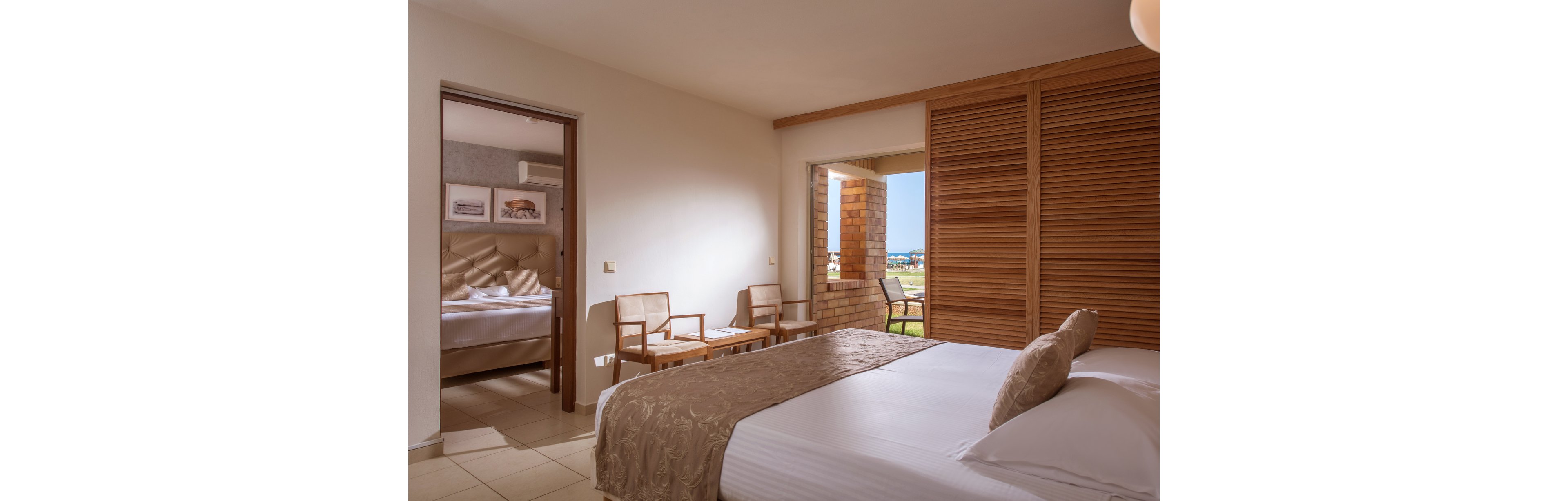 A sunlit hotel bedroom features a neatly made bed with neutral bedding and pillows. Sliding wooden shutters open to a terrace with chairs and a view of an outdoor landscape. In the adjoining room, another bed and framed wall art are visible through an open doorway. The overall scene feels calm, tidy, and inviting, with warm tones and soft natural light.
