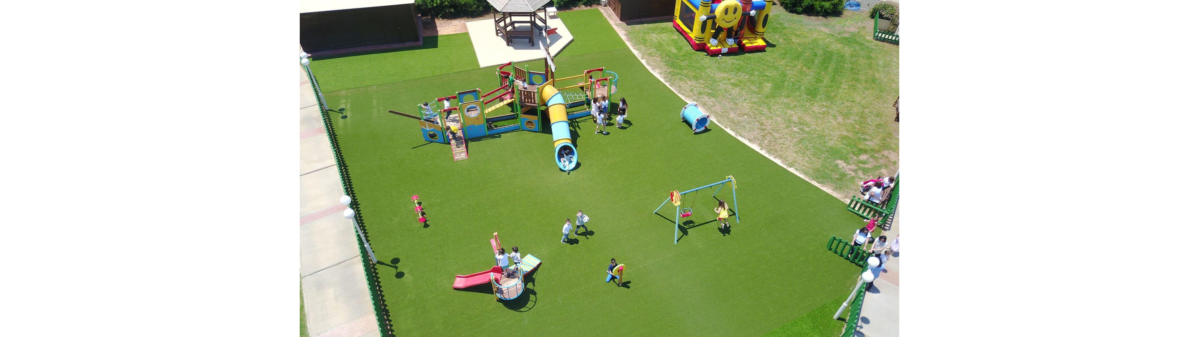 An aerial view of a vibrant playground featuring slides, swings, and climbing structures on a green artificial turf. Children are actively playing and enjoying various activities in the open space. A bouncy castle and gazebo are visible in the background, adding to the lively atmosphere.