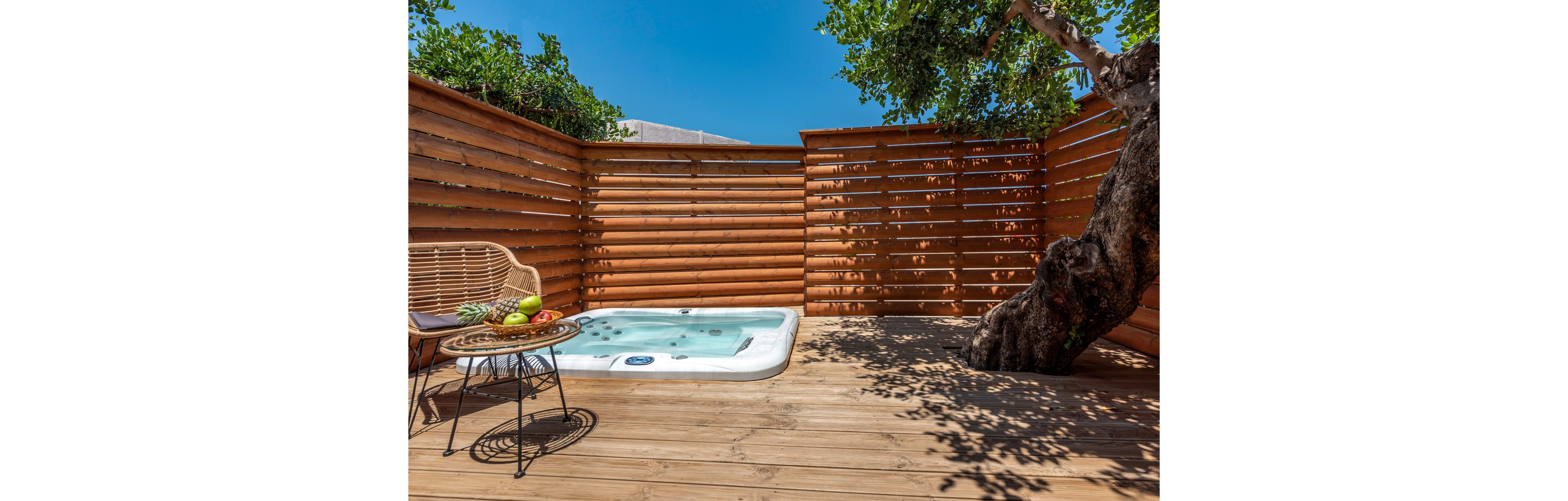 A compact outdoor hot tub set on a wooden deck surrounded by a privacy fence. The setting includes a wicker chair and a small table with fresh fruits, creating a relaxing atmosphere. A large tree adds natural charm to the scene under a clear blue sky.