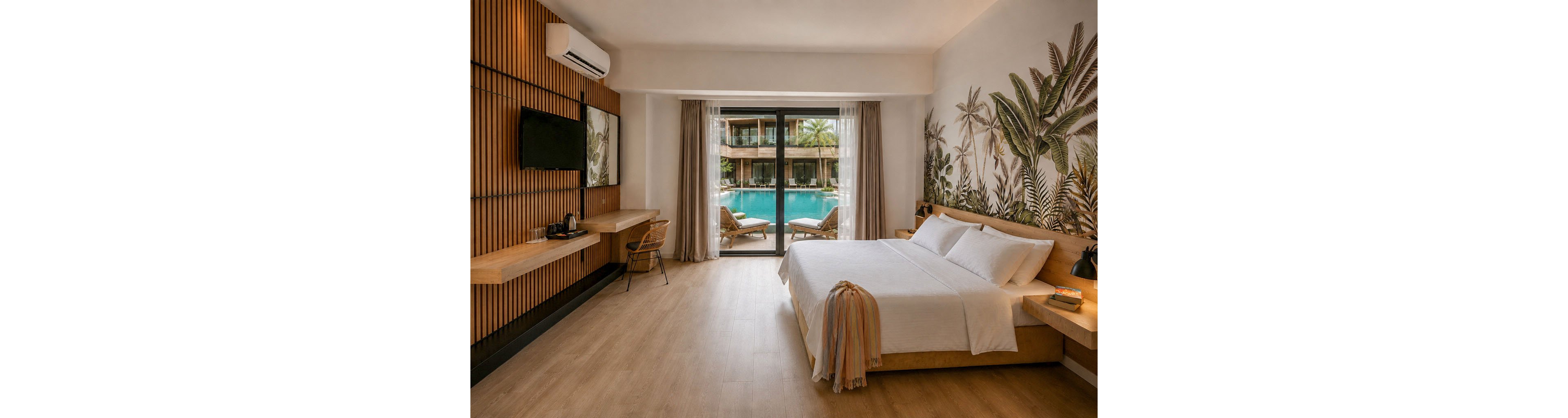 A contemporary hotel bedroom features a large bed with white linens and a light throw draped over the foot. Floor-to-ceiling glass doors open to a balcony overlooking a swimming pool and sun loungers. The interior includes warm wood paneling, a wall-mounted television, and a minimalist desk with a kettle and cups. A botanical mural decorates the wall behind the bed, enhancing the tropical resort atmosphere.