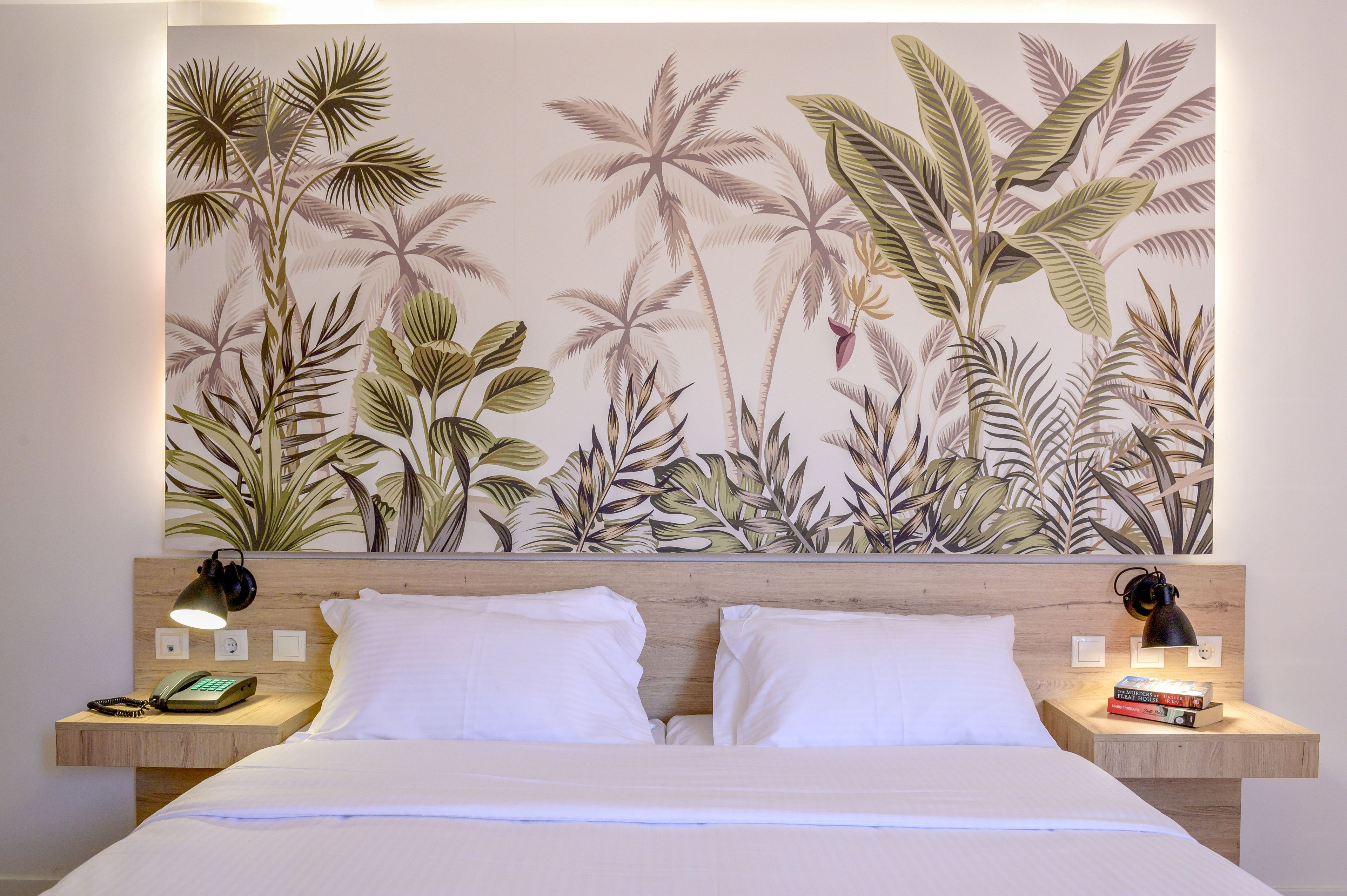 A neatly made double bed with white linens sits against a light wood headboard in a contemporary hotel room. Above the bed, a large mural features tropical palm trees and lush foliage in muted green and beige tones. Two black bedside lamps provide warm lighting on either side, with a telephone and books placed on the floating nightstands. The overall atmosphere is clean, inviting, and stylish, combining minimal design with nature-inspired decor.