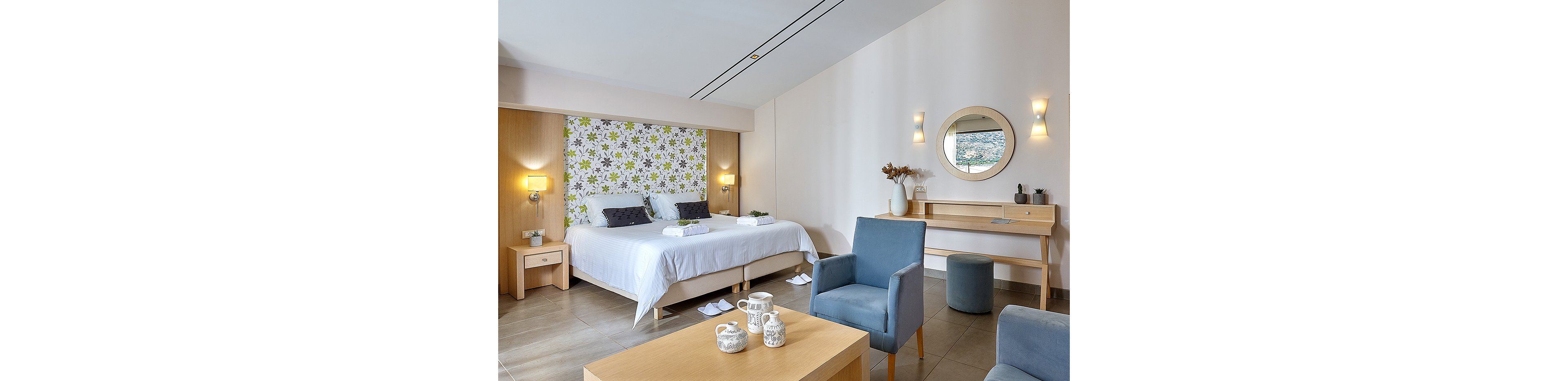 Contemporary hotel bedroom featuring a double bed with white linens and floral patterned headboard. The room includes bedside tables, wall-mounted lamps, and folded towels placed neatly on the bed. A cozy seating area with blue armchairs and a wooden coffee table holds decorative ceramics. A minimalist wooden desk with round mirror, stool, and vases completes the light, airy interior design.