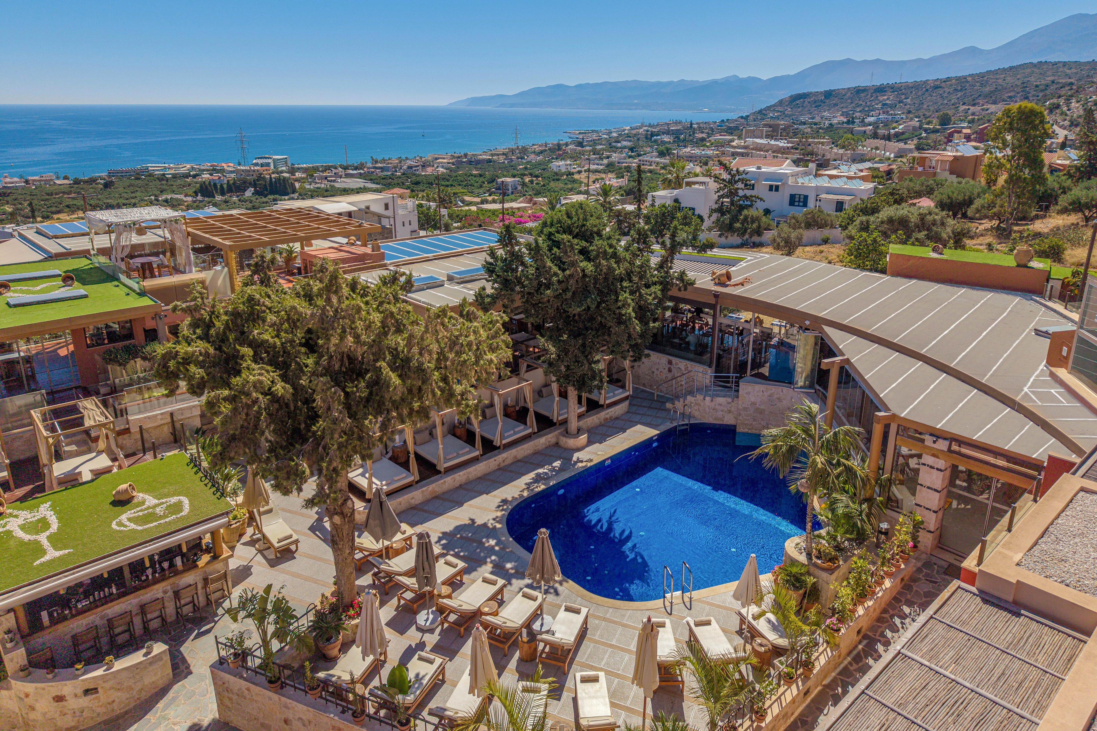 A hilltop resort features a curved blue swimming pool surrounded by sun loungers and umbrellas. Outdoor dining and bar areas with wooden structures and greenery frame the pool. Beyond the resort, a coastal town stretches toward the sea with distant mountains on the horizon. The bright daylight and clear sky create a warm, inviting vacation atmosphere.