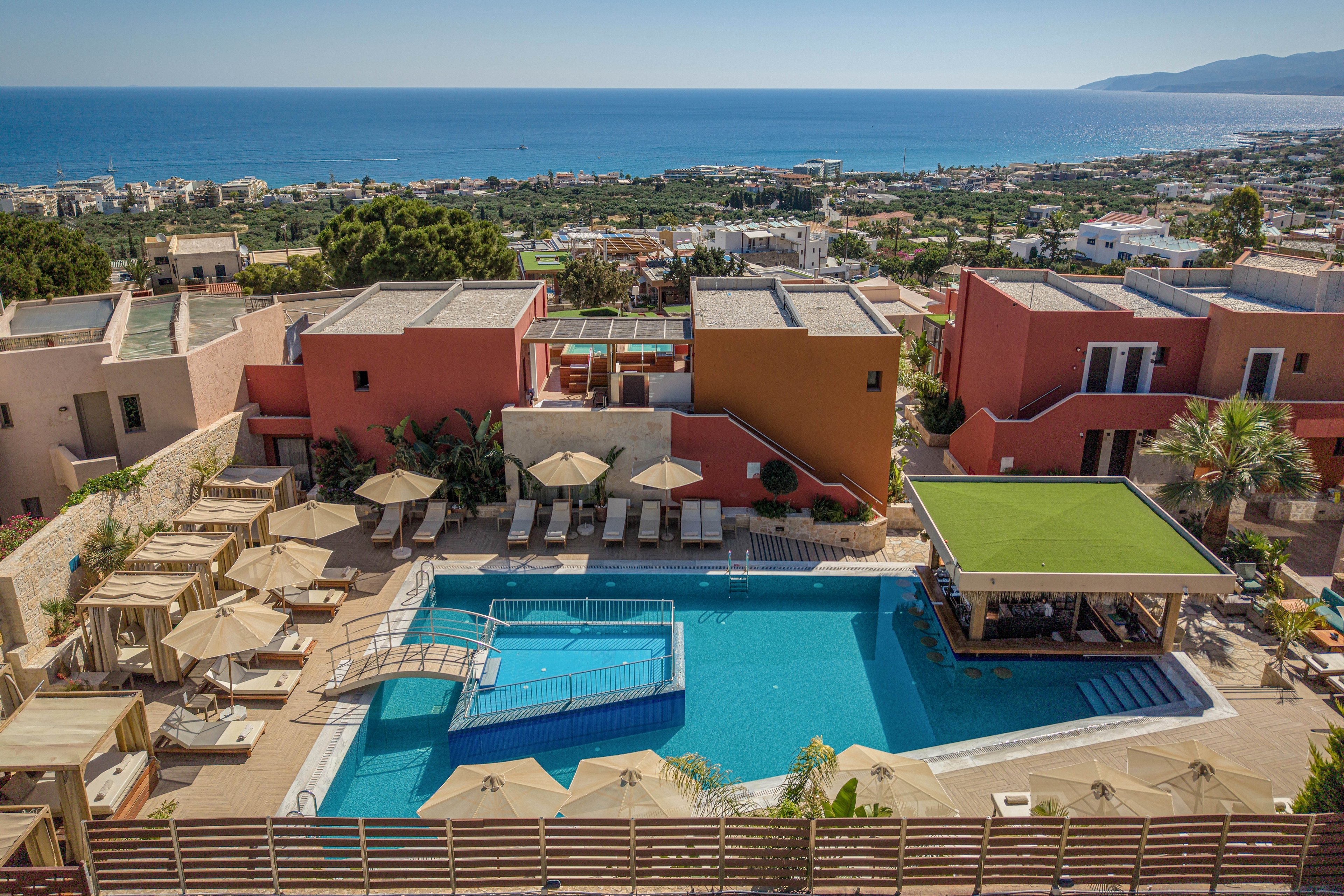 A large outdoor swimming pool sits at the center of a modern resort complex with colorful buildings and sun loungers. Beige umbrellas and cabanas surround the pool, creating a relaxed holiday atmosphere. A small bridge crosses part of the pool, and a covered bar or lounge area with a green roof is visible on one side. In the background, a coastal town stretches toward the sea, with blue water and distant mountains completing the scenic view.