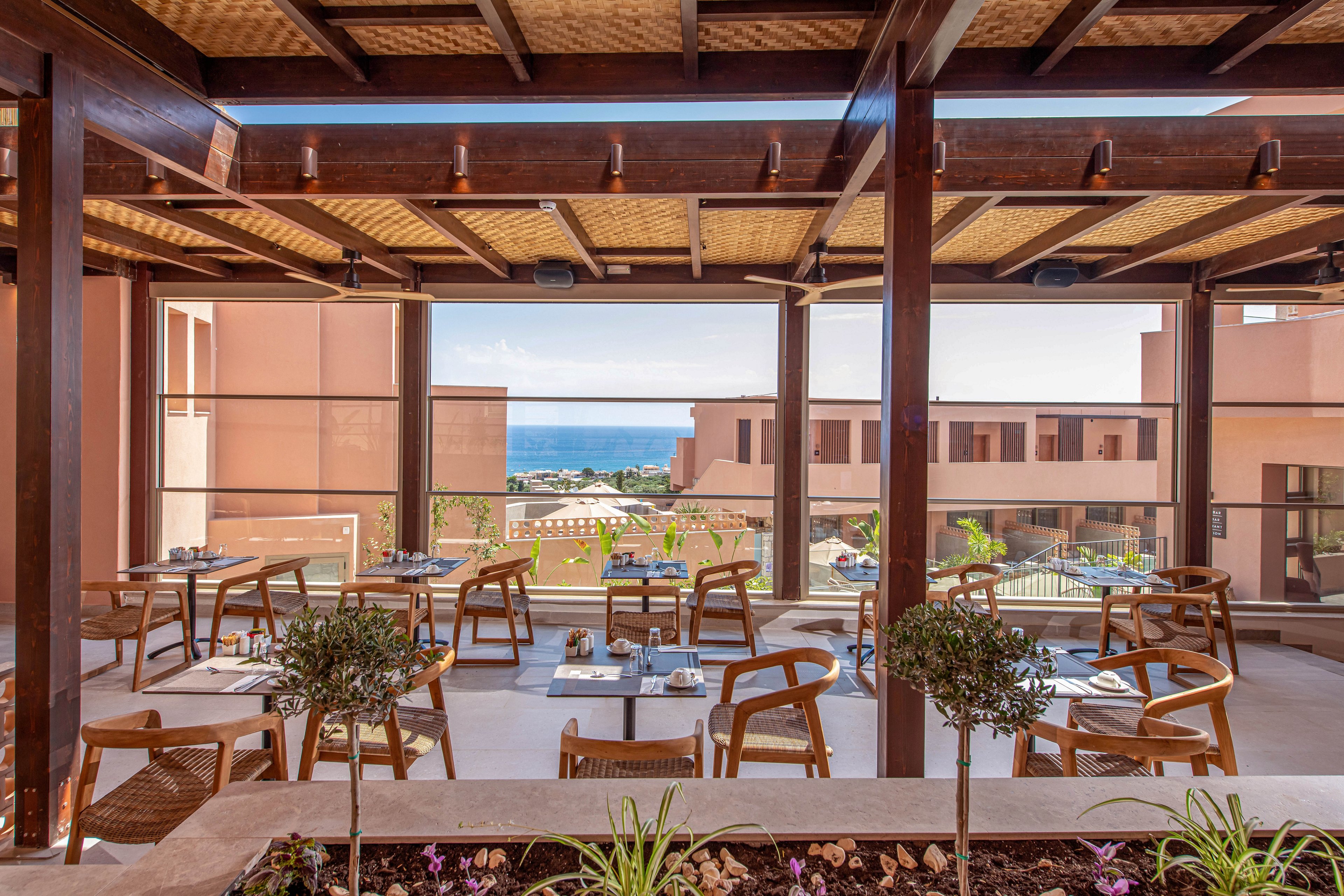 An open-air terrace restaurant features wooden tables and chairs arranged for casual dining. The setting overlooks a modern resort complex with peach-colored buildings and landscaped grounds. Large wooden beams and a woven ceiling frame the view toward the distant blue sea and sky. Potted plants and a small garden bed add greenery and a relaxed vacation atmosphere.