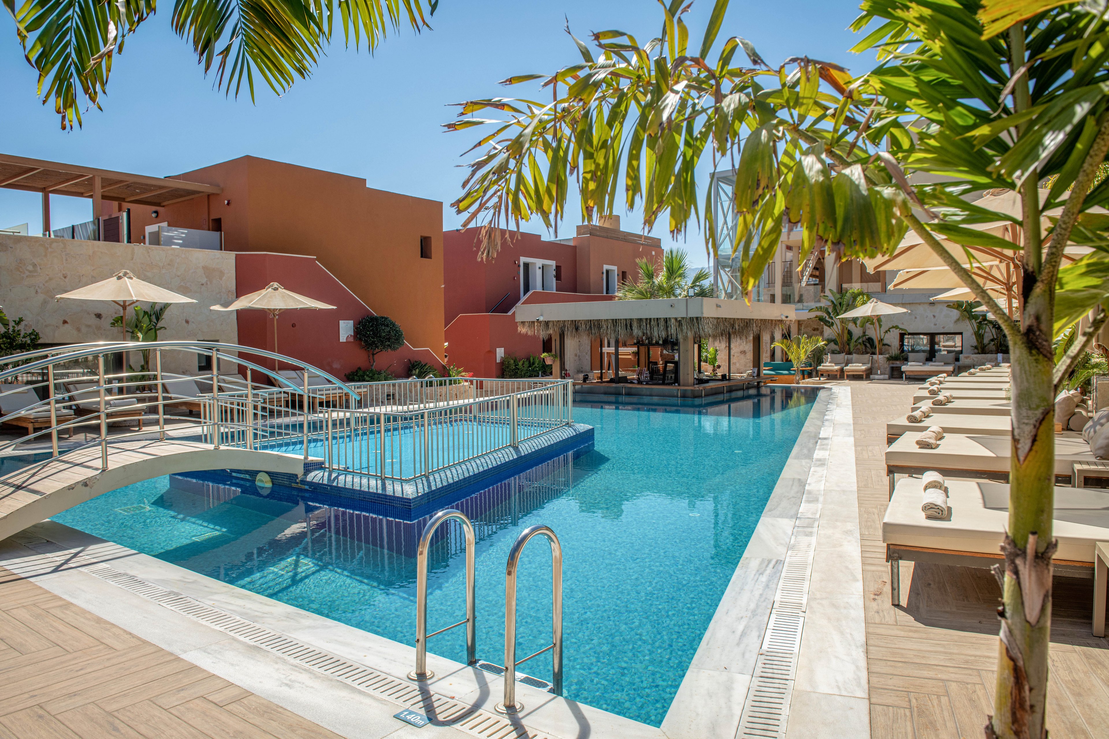 Outdoor resort swimming pool with a small white bridge crossing over the water. Sun loungers with rolled towels line the deck under beige umbrellas, suggesting a relaxing vacation setting. Modern low-rise buildings in warm terracotta tones surround the pool area. Palm trees and clear blue sky create a bright, tropical atmosphere.