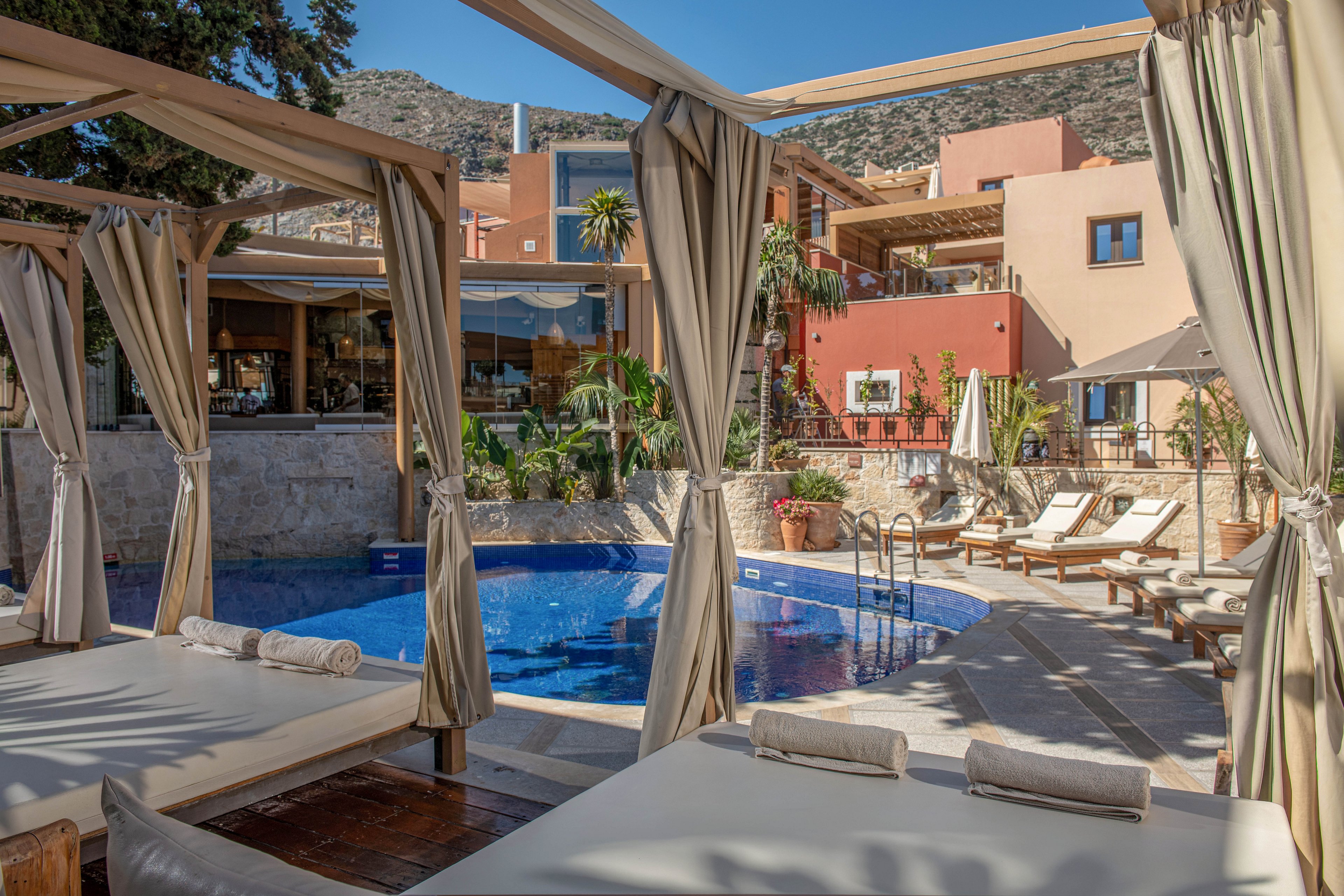 Outdoor hotel pool area with cushioned cabana beds and sun loungers arranged around a small blue swimming pool. Neutral fabric curtains frame the cabanas, creating shaded relaxation spaces in the foreground. Warm-toned buildings with balconies, potted plants, and palm trees surround the courtyard, suggesting a Mediterranean or resort setting. The scene is calm and inviting, with clear daylight and soft shadows emphasizing a tranquil vacation atmosphere.