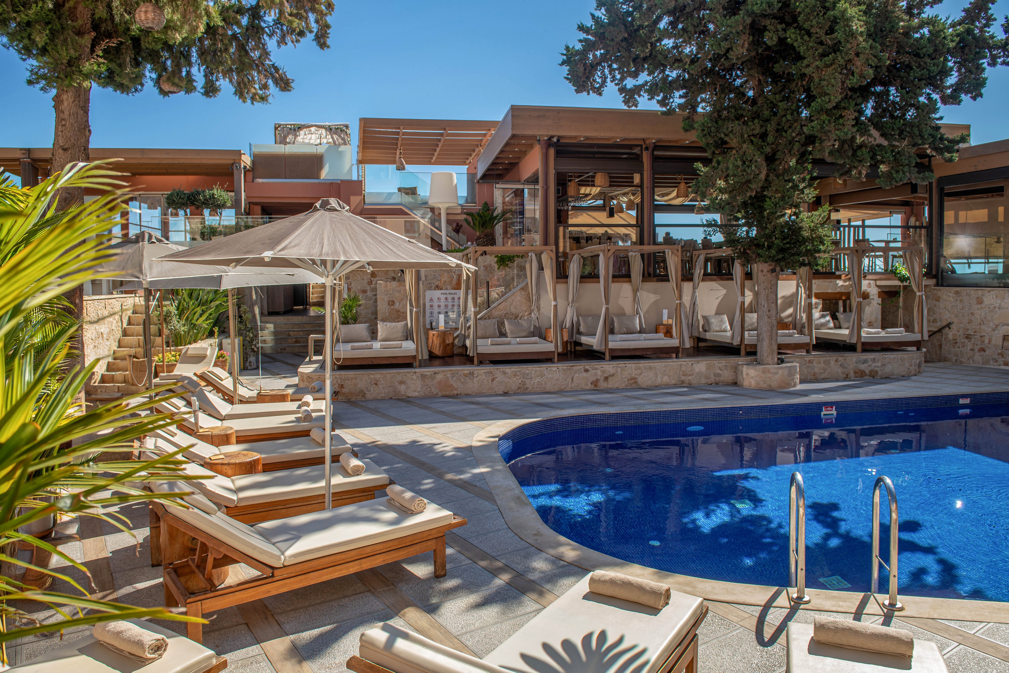Outdoor resort pool area with wooden sun loungers and beige cushions arranged around a curved blue swimming pool. Large umbrellas provide shade, and cabana-style daybeds line the terrace behind the pool. The setting is bright and sunny with clear blue sky, stone walls, and lush greenery adding a Mediterranean feel. Stainless steel pool ladders and neatly placed towels emphasize a relaxed, upscale vacation atmosphere.