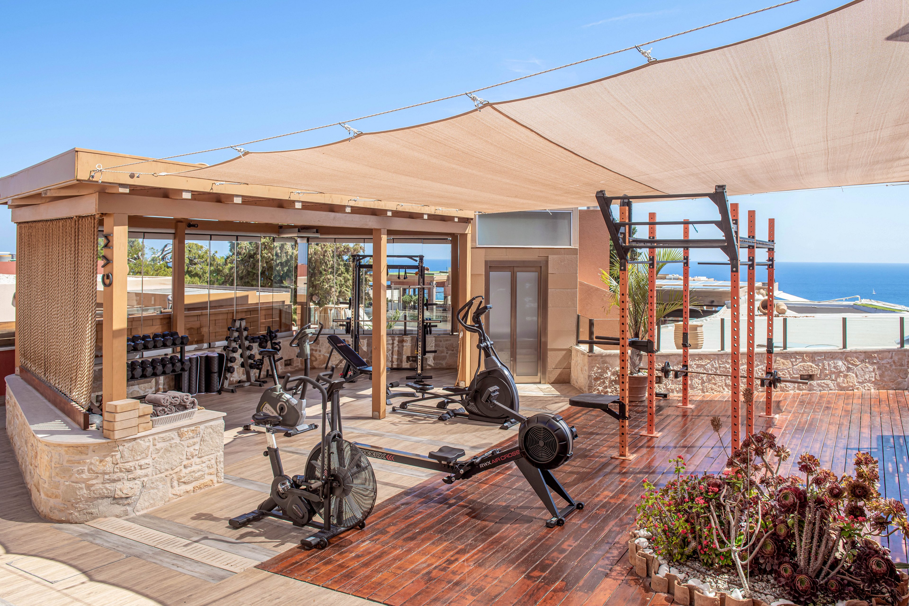An open-air rooftop gym features various fitness equipment under a light fabric canopy. Cardio machines and free weights are arranged on a wooden deck beside a glass-walled workout area. A functional training rig stands near potted plants, adding a touch of greenery to the space. In the background, the blue sea and clear sky create a bright, relaxing atmosphere for exercise.