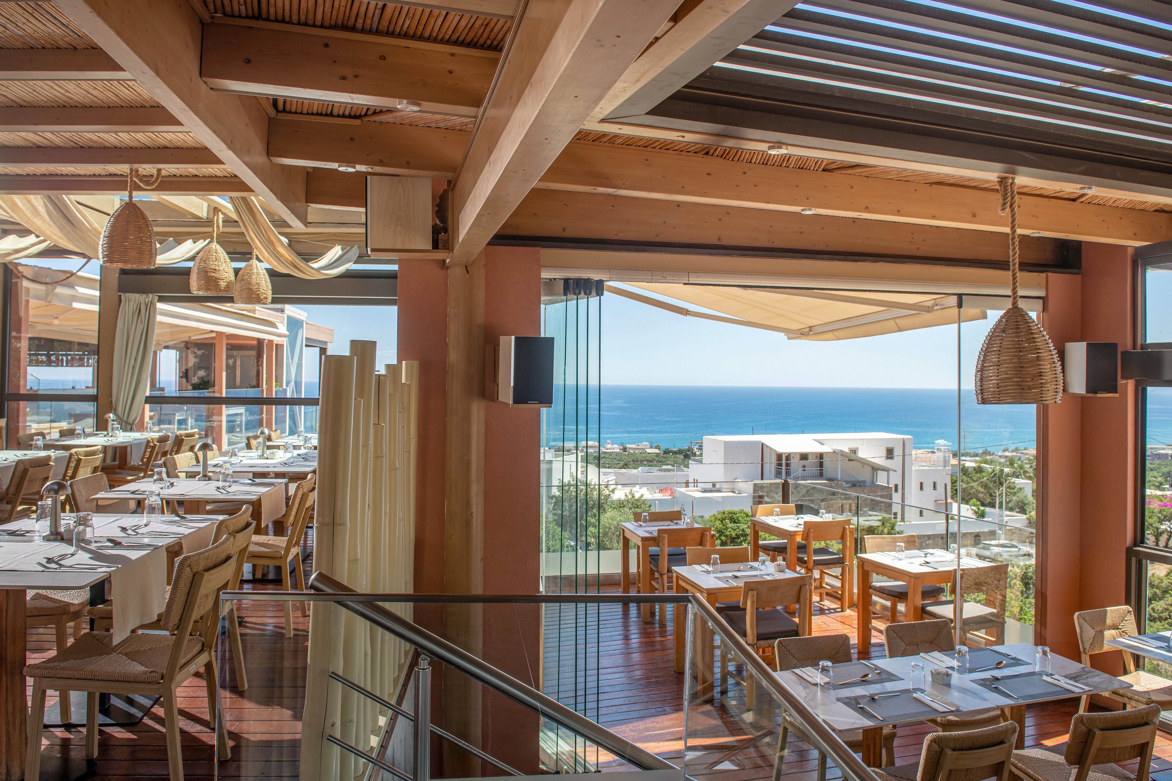 Bright, modern restaurant interior overlooks a coastal town and blue ocean horizon. Wooden tables and chairs are neatly arranged with white tablecloths, glassware, and cutlery ready for guests. Large windows and open-air terraces allow natural light to flood the space, complemented by slatted wooden ceilings and hanging wicker lamps. The atmosphere feels relaxed and inviting, ideal for leisurely dining with scenic sea views.