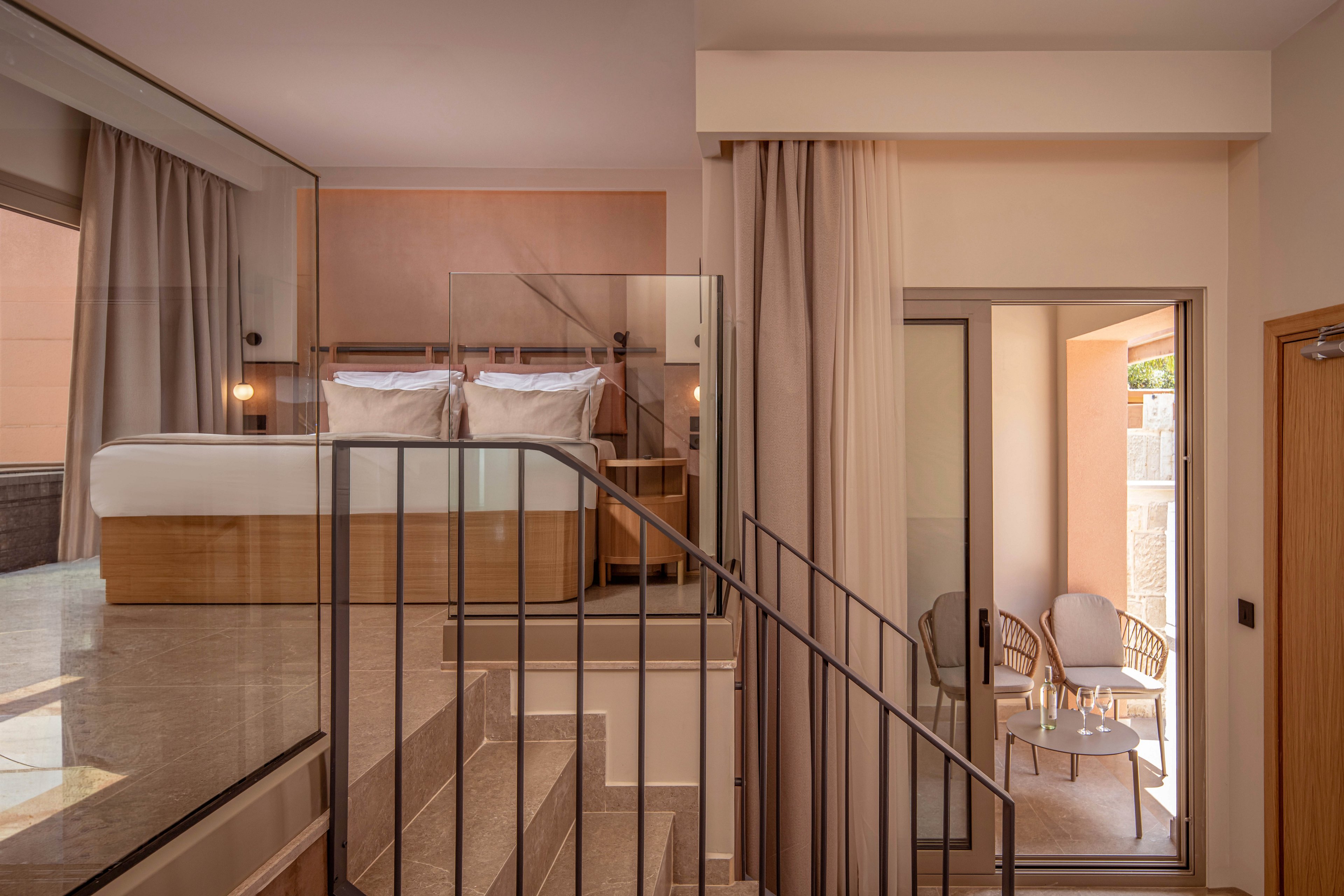 Contemporary split-level interior features a raised bed area with two white pillows and neutral bedding. A staircase with metal railing and glass panels connects the sleeping space to the lower level. Floor-to-ceiling curtains frame a sliding glass door that opens to a small balcony. Outside, two woven chairs and a round table create a cozy seating nook in warm natural light.