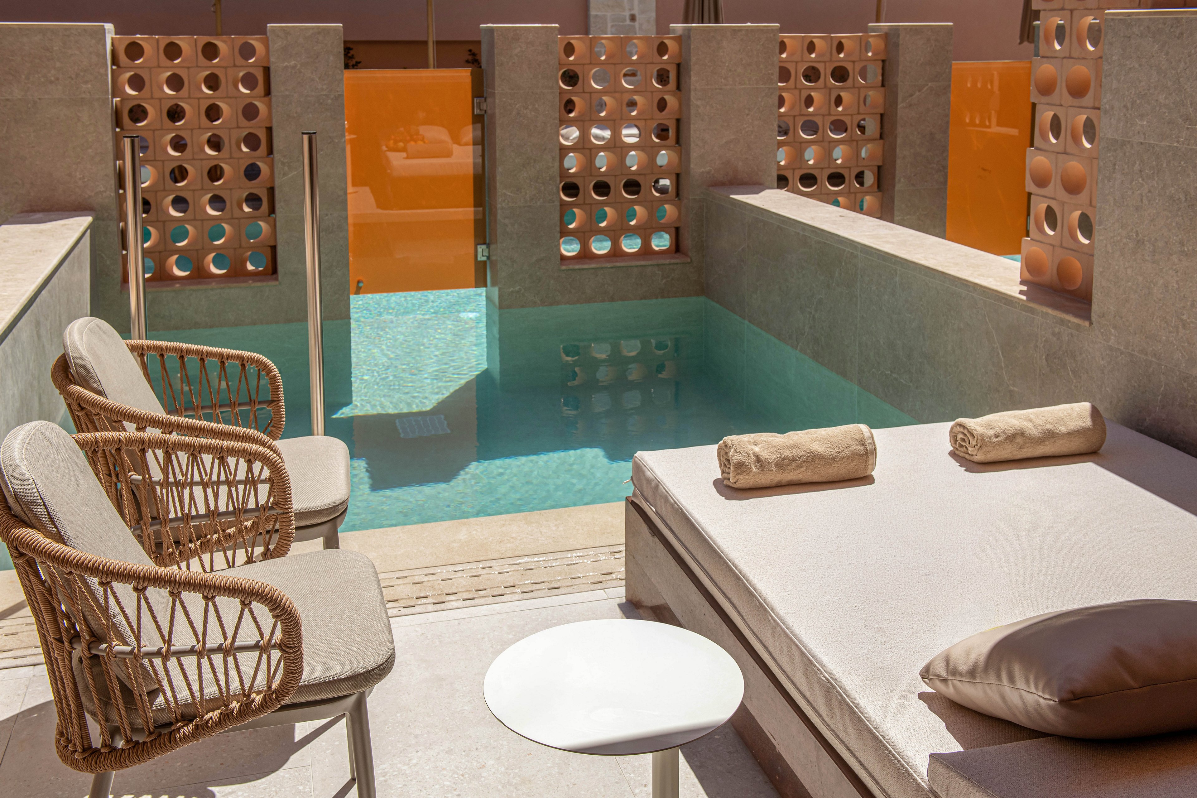 A small private plunge pool is surrounded by modern stone walls and warm-toned decorative screens. A cushioned sunbed with rolled towels sits beside the water, creating a relaxing resort atmosphere. A round side table holds a bottle of white wine and two empty wine glasses. A large plate of fresh fruit, including a pineapple, apples, oranges, and peaches, adds vibrant color to the serene outdoor setting.