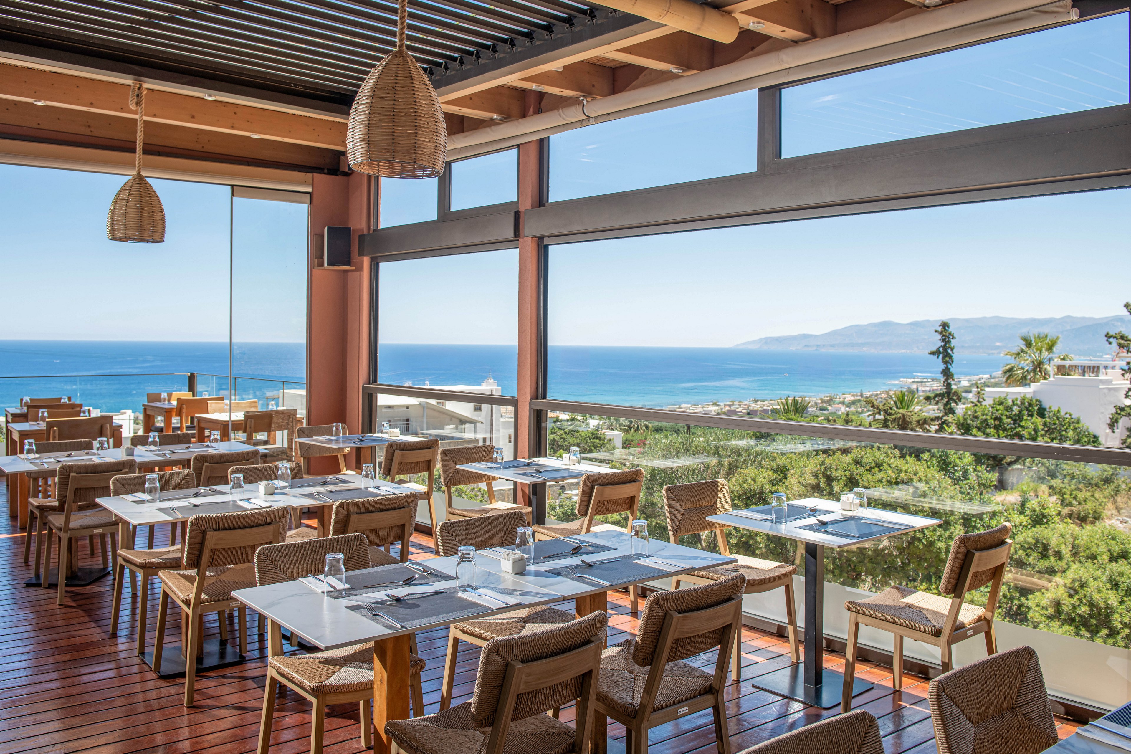 A modern open-air restaurant overlooks a bright blue ocean and distant mountains. Wooden tables and chairs are neatly arranged with glassware and place settings, ready for guests. Large floor-to-ceiling windows and railings frame the coastal landscape and lush greenery below. Warm natural materials and soft daylight create a relaxed, inviting atmosphere for dining with a view.