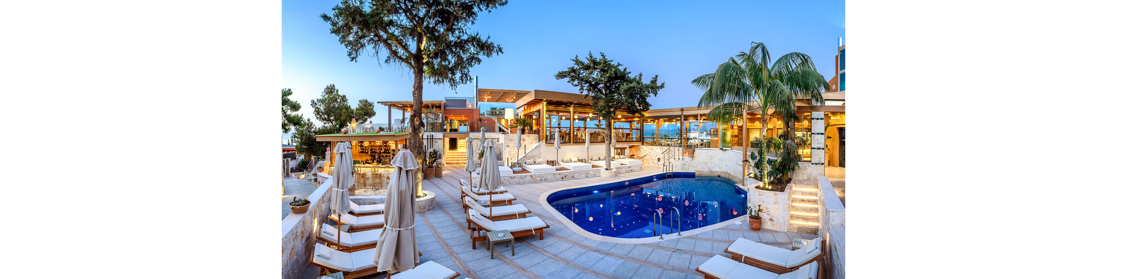 A contemporary resort pool area is shown at dusk with lounge chairs neatly arranged around a curved blue swimming pool. Beige umbrellas are folded beside the sunbeds, and warm lights illuminate the stone terraces and stairways. The open-air restaurant and bar in the background feature large windows and wooden accents, creating an inviting atmosphere. Tall trees and palm plants frame the scene, while the clear evening sky adds to the tranquil vacation setting.