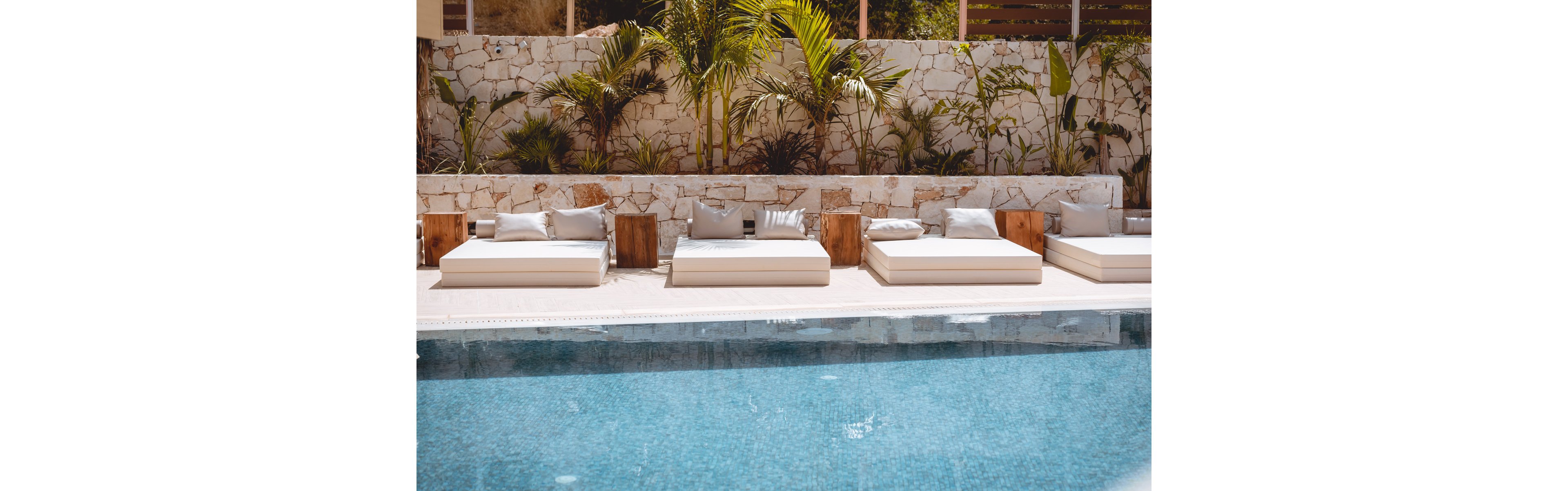 A row of cushioned lounge beds is arranged neatly beside a clear blue swimming pool. The setting features a stone wall backdrop with lush green tropical plants. Neutral-toned pillows and wooden side tables create a modern, minimalist aesthetic. Bright daylight enhances the tranquil, resort-like atmosphere.