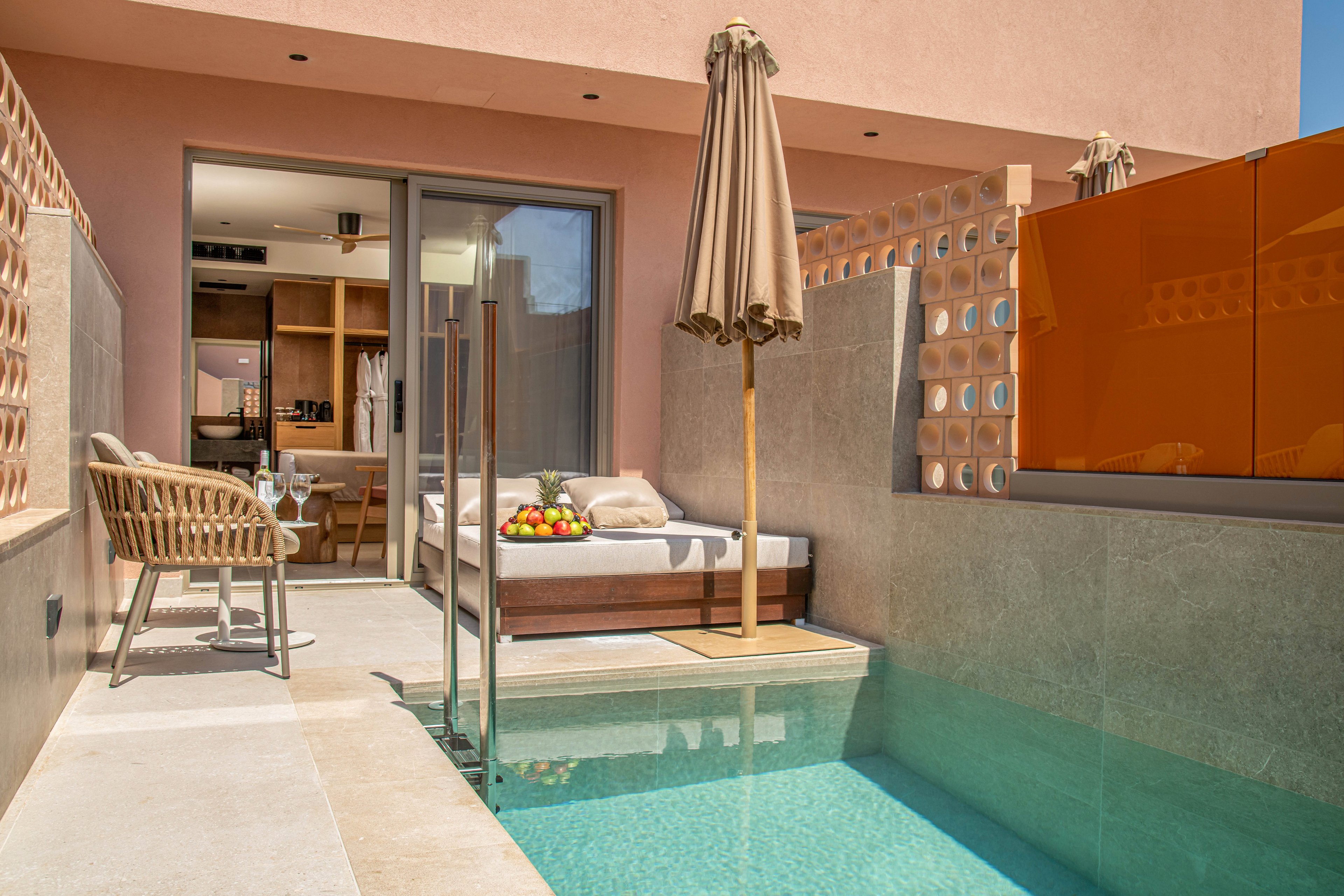 A contemporary private terrace features a small turquoise pool beside a cushioned sunbed topped with a fruit platter. A wicker chair and table with two wine glasses create an inviting seating area under a tall beige umbrella. Sliding glass doors open into a stylish interior with wooden cabinetry and soft lighting. Warm neutral tones and clean lines give the outdoor space a relaxed, luxurious resort feel.