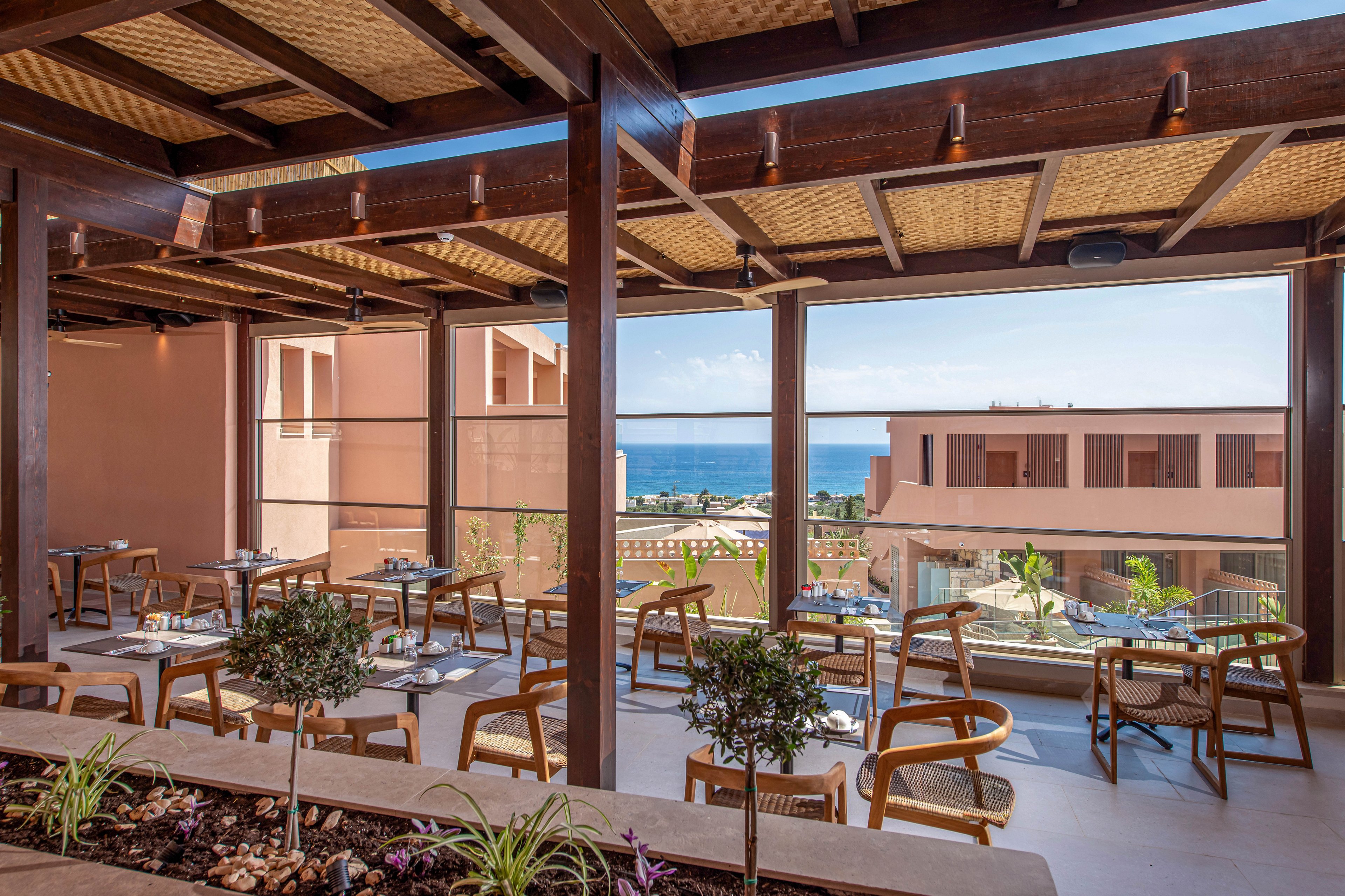 An open-air terrace restaurant features wooden tables and chairs arranged neatly under a pergola roof. Large windows frame a view of modern resort buildings and the distant blue sea. Potted plants and a small planter with stones and greenery add a natural touch to the contemporary setting. Warm earth tones and natural materials create a relaxed, inviting atmosphere for outdoor dining.