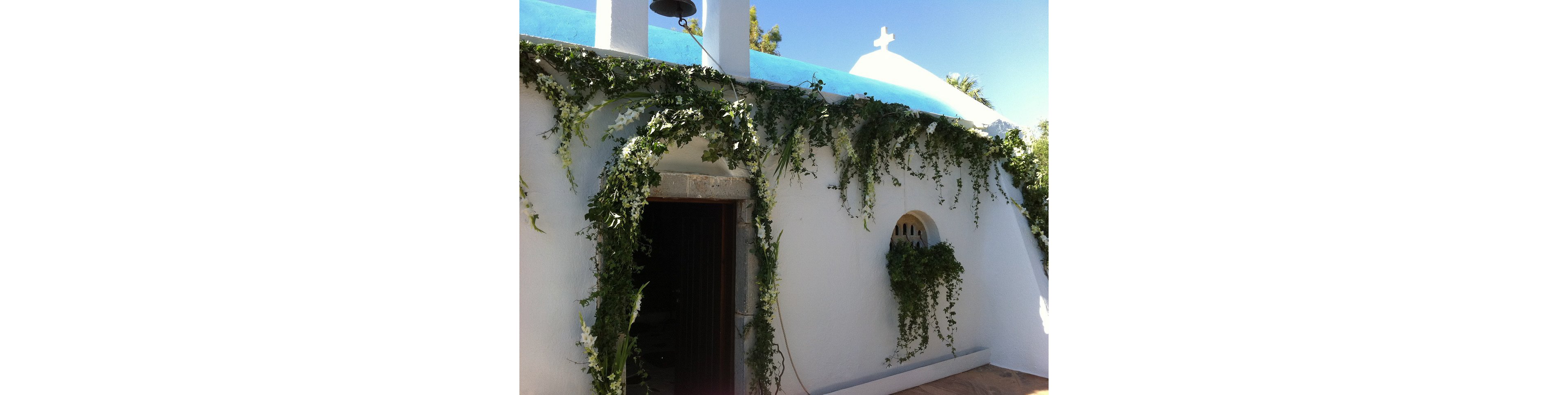 A small whitewashed chapel is adorned with lush green garlands draped along its exterior walls. The building features a simple arched doorway and a small round window framed by foliage. A blue roofline and dome with a white cross stand out against the clear sky. Bright sunlight emphasizes the clean lines and Mediterranean feel of the scene.