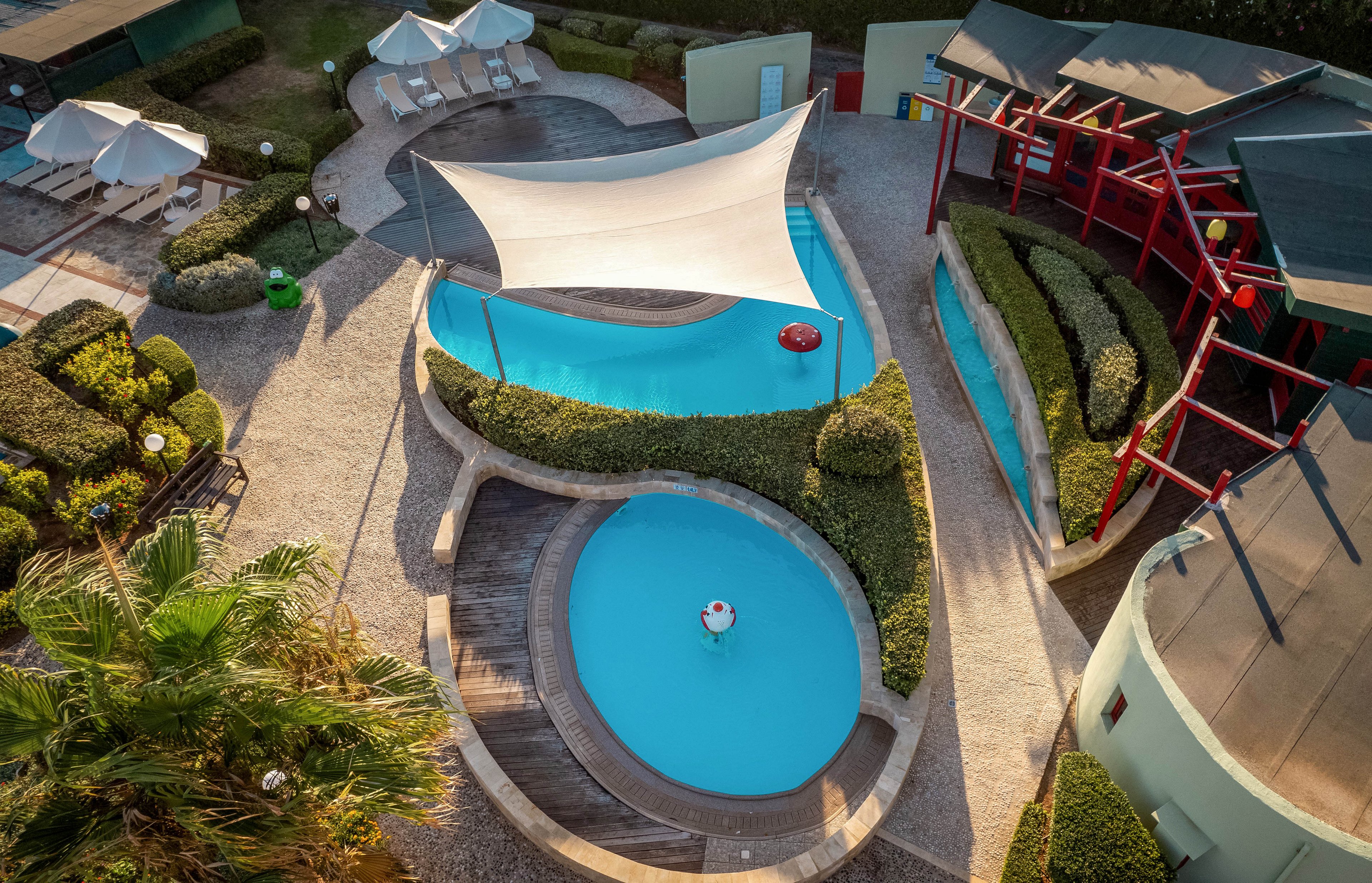 This image shows a contemporary outdoor swimming pool complex photographed from above. The scene features two uniquely shaped blue pools, surrounded by curved walkways, manicured hedges, and sun loungers with white umbrellas. A large white shade sail covers part of the main pool, and the area is accented by red architectural elements and lush greenery. No visible text or numbers are present in the image.