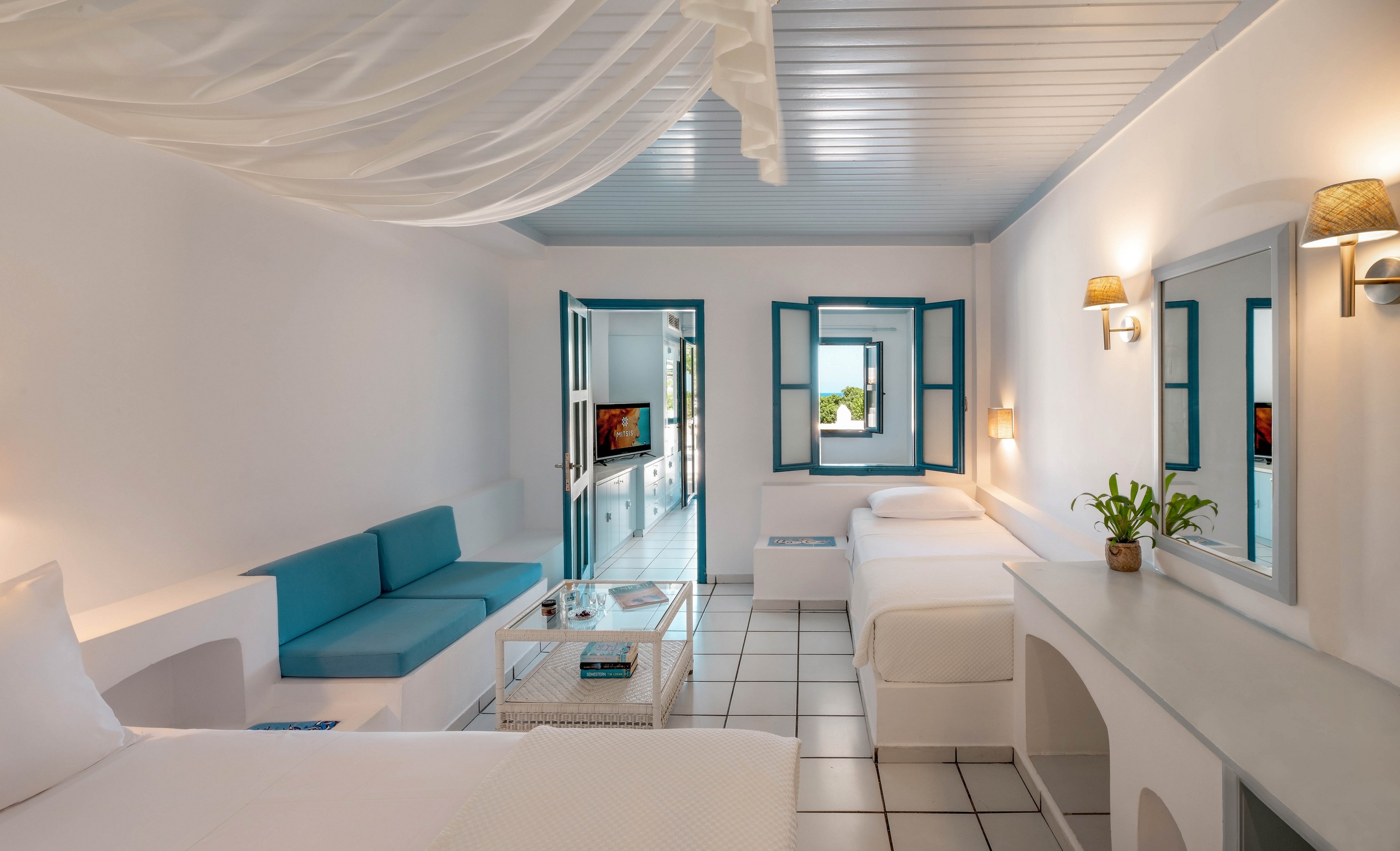 A spacious hotel room features white walls, blue window frames, and minimalist furnishings. The setting includes two beds, a small sofa, a coffee table, and a vanity with a mirror, all arranged in a clean, airy layout. Natural light streams in through open windows, highlighting the serene and inviting atmosphere. The room is decorated with potted plants and soft lighting, creating a relaxing mood. No visible text or numeric values are present in the image.