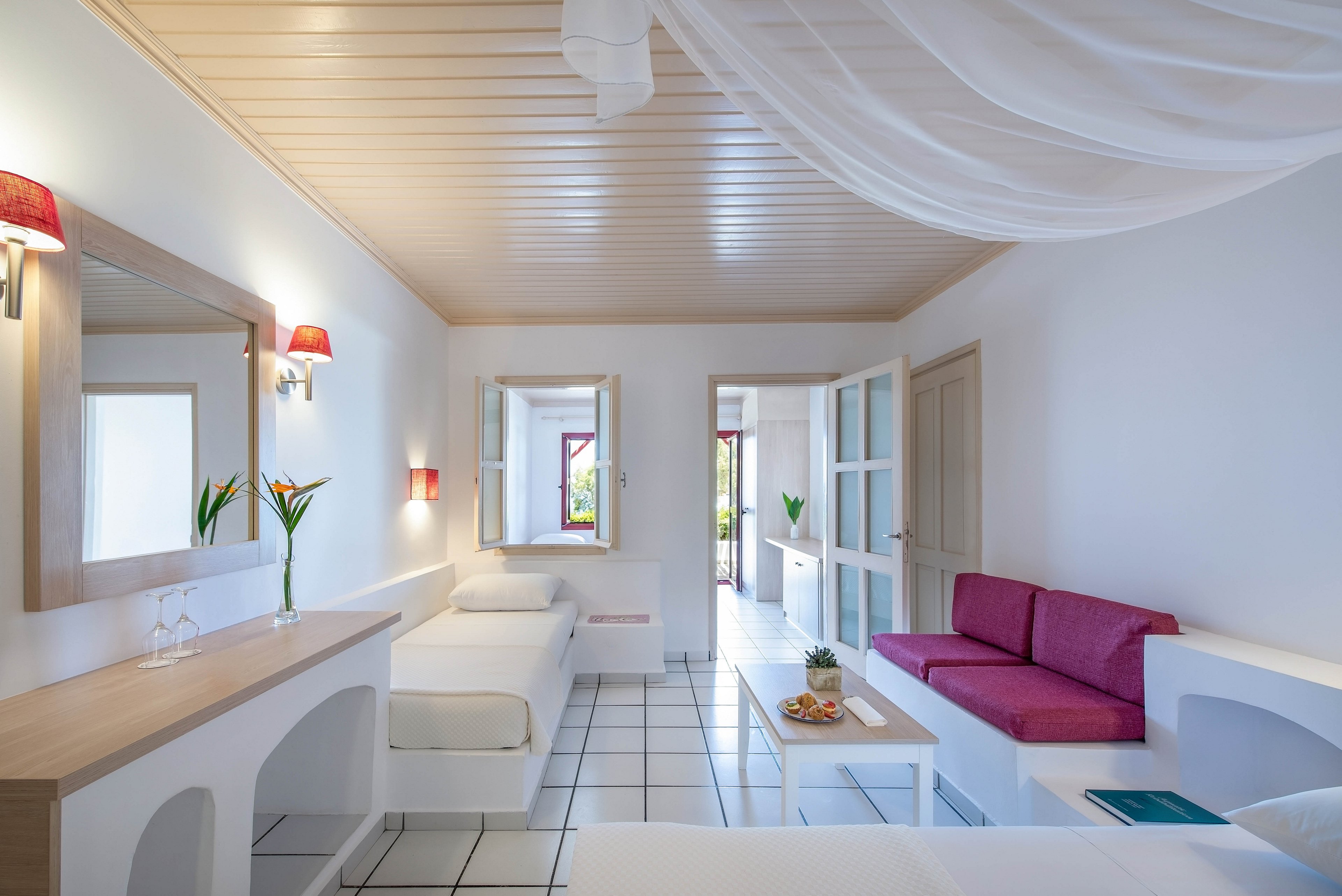 A spacious hotel room features a contemporary design with white walls, tiled floors, and a light wood ceiling. The room includes a pink sofa, two single beds, and a table with snacks and a bottle of rosé wine. Decorative touches include tropical flowers in vases and soft lighting from wall sconces. The atmosphere is airy and inviting, with natural light streaming in from the open doorway.