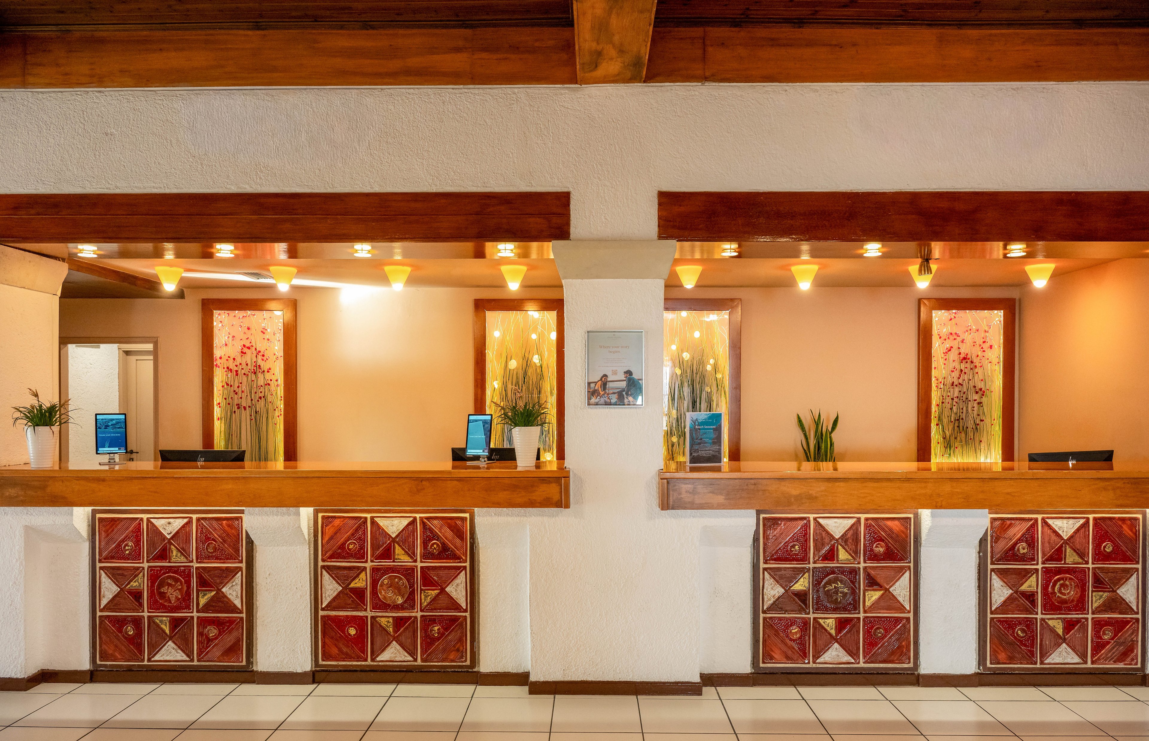 Three receptionists stand behind a wooden hotel front desk, each at their own station with computers and potted plants. The setting is warmly lit with decorative wall art and geometric tile patterns on the desk. The environment appears welcoming and professional, with a neutral color palette and modern decor elements.
