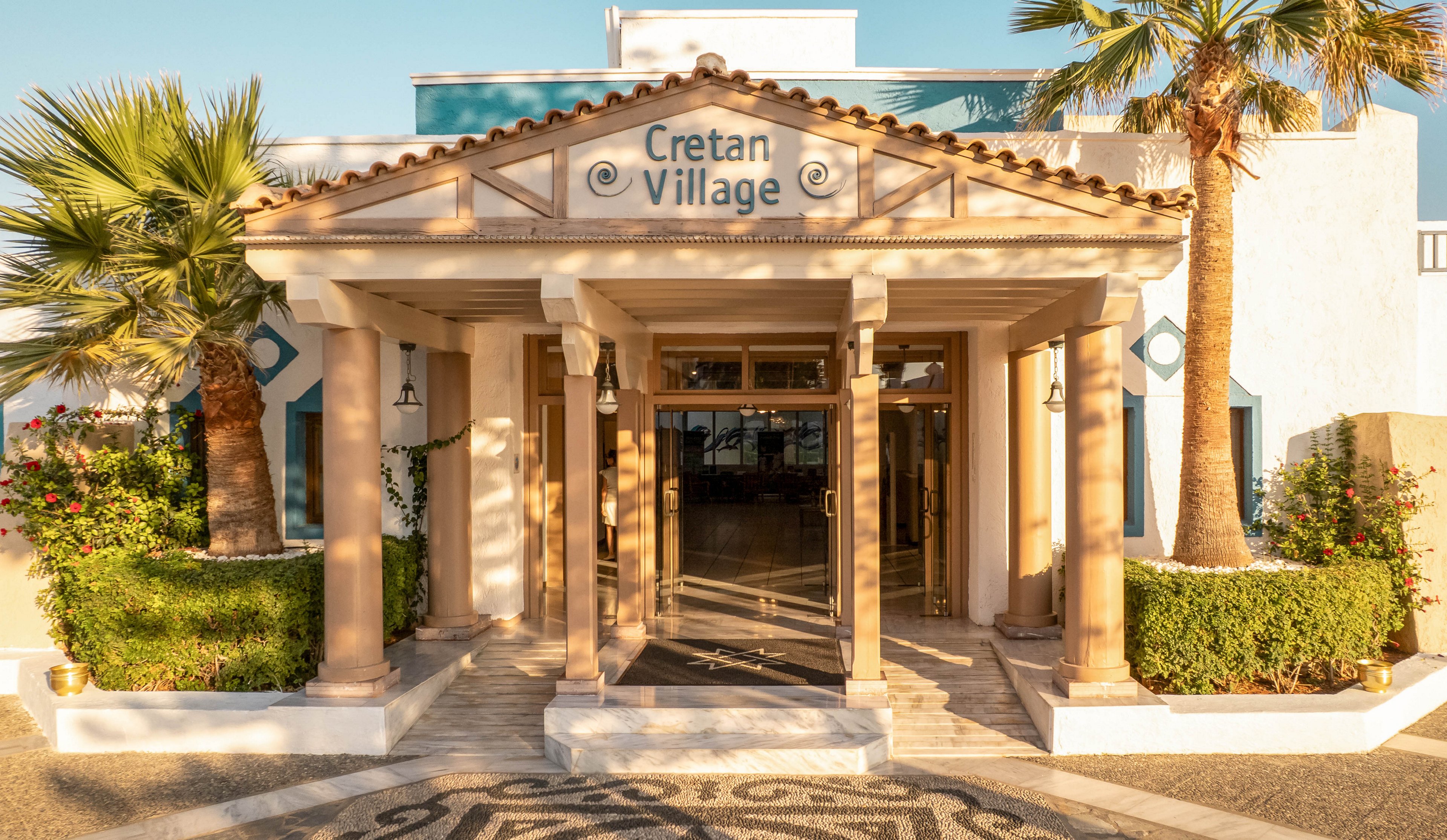 The image shows the main entrance of a building labeled 'Cretan Village,' featuring classical columns and a tiled mosaic walkway. The setting is outdoors, with lush greenery and palm trees framing the entrance, suggesting a Mediterranean or resort ambiance. The lighting is warm and golden, indicating late afternoon or early evening. The architectural style is traditional, with a welcoming and serene mood.
