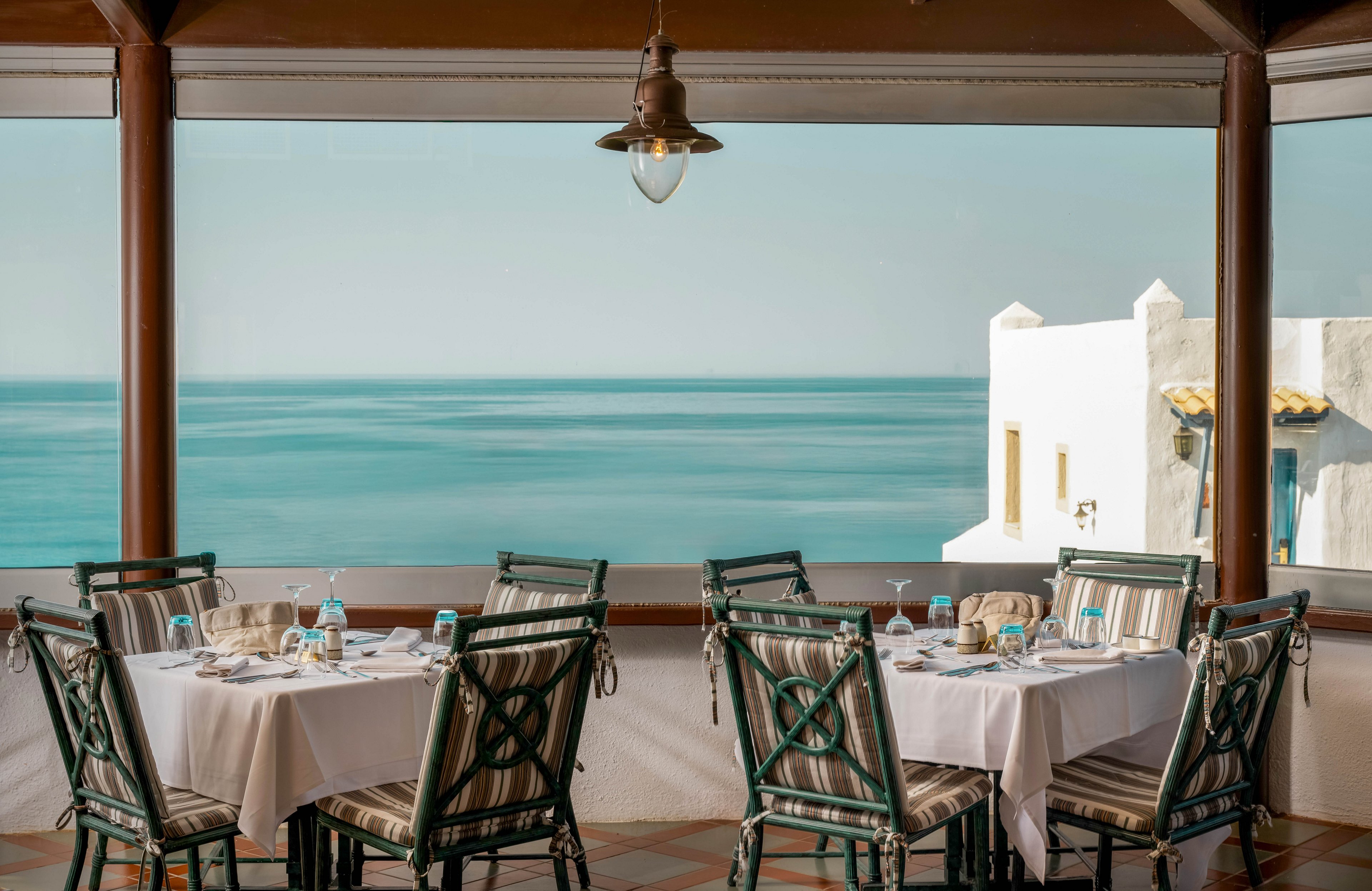 The image features a restaurant terrace overlooking a calm blue ocean, with neatly set tables and striped chairs. The setting is tranquil and sunlit, evoking a relaxed Mediterranean mood. Whitewashed architecture is visible in the background, enhancing the coastal ambiance. No visible text or numeric values are present in the scene.