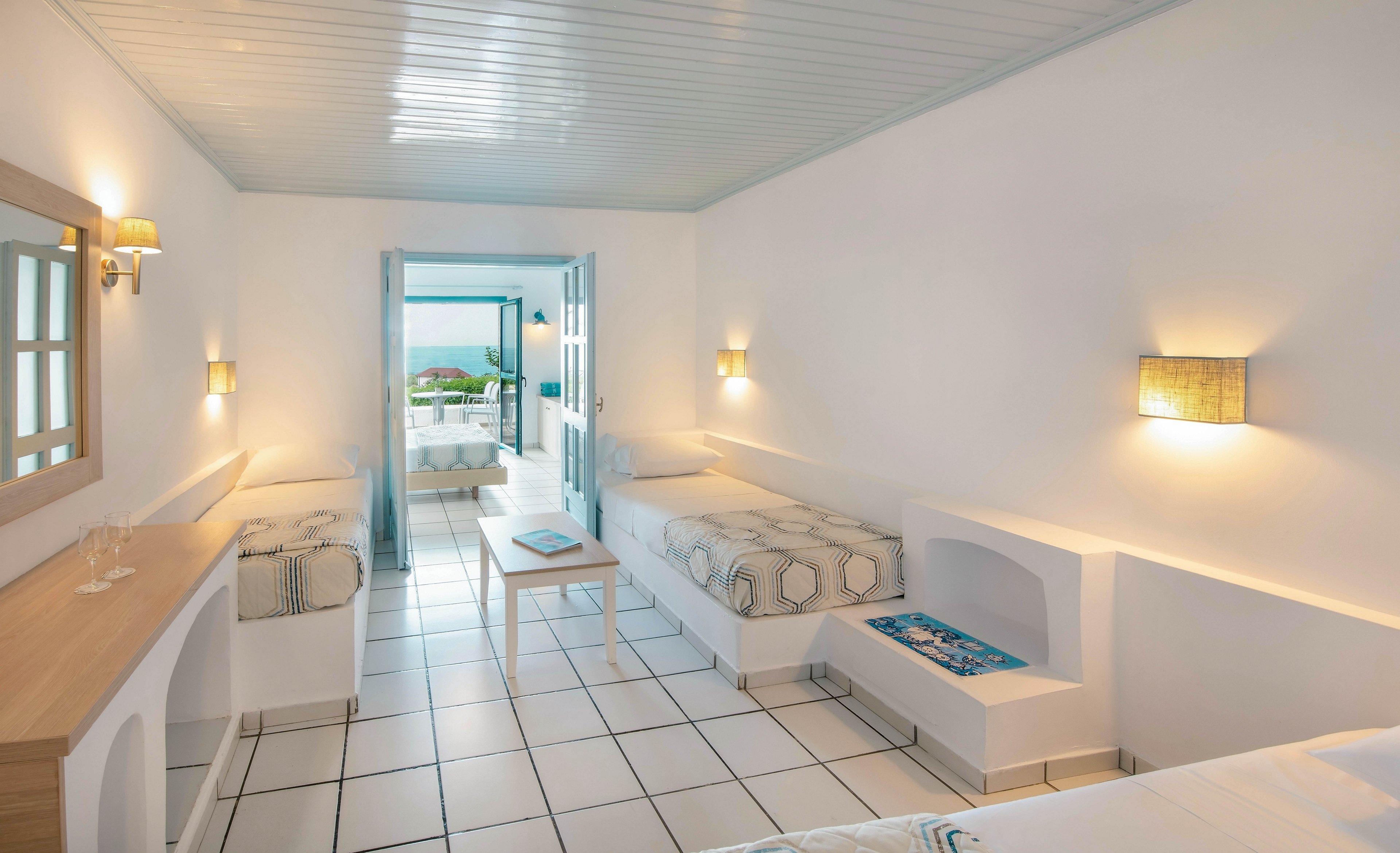 A spacious, white-walled hotel room features minimalist built-in beds and furniture, accented with soft lighting and geometric patterned linens. The room opens to a terrace with a view of the ocean and distant landscape. A bottle and glasses are arranged on the wooden counter, and the overall palette is light and airy, emphasizing relaxation and tranquility.