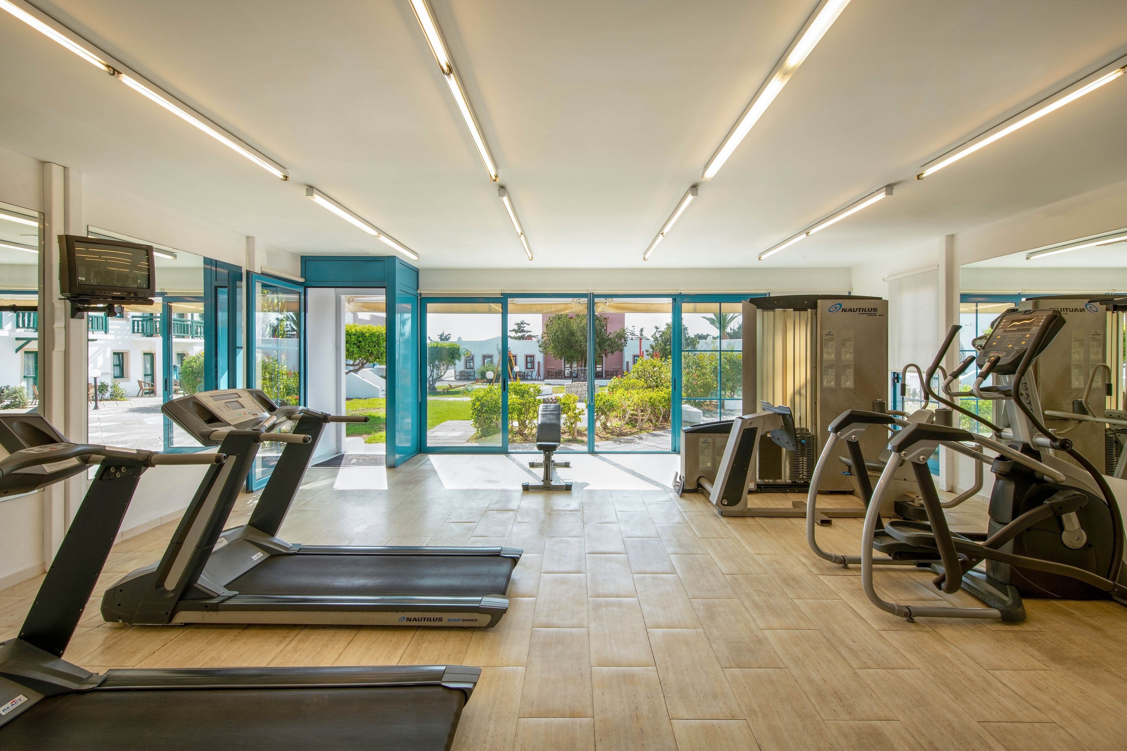 A bright, spacious gym features treadmills, elliptical machines, and strength training equipment. Large windows provide natural light and a view of a green outdoor area. The interior has a clean, minimalist design with light wood flooring and white walls. No people are present, emphasizing the equipment and open space.