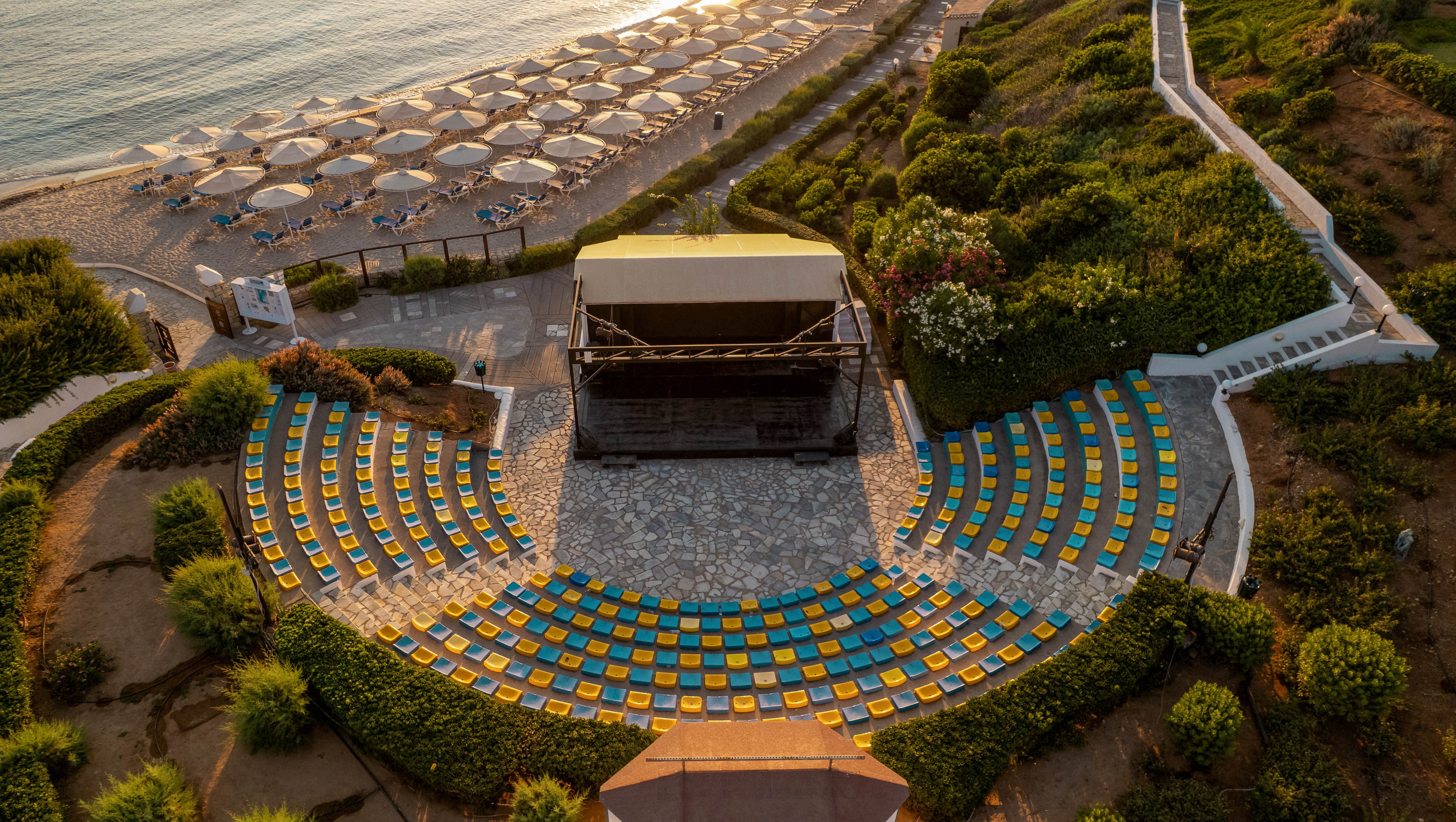 Aerial view of a semi-circular outdoor amphitheater with rows of colorful seats facing a central stage. The venue is set on a hillside with lush greenery, adjacent to a sandy beach lined with umbrellas and lounge chairs. The scene is bathed in warm, golden sunlight, creating a tranquil and inviting atmosphere.