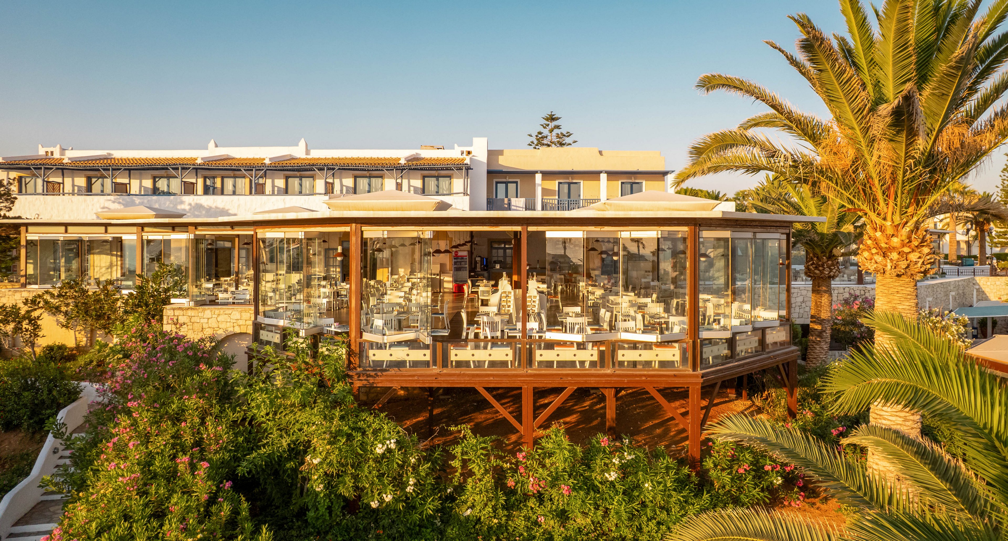 A contemporary restaurant with floor-to-ceiling glass windows is elevated above a vibrant garden filled with greenery and palm trees. The setting is outdoors, bathed in warm golden sunlight, suggesting a relaxed and inviting mood. The restaurant interior features neatly arranged tables and chairs, with no visible text or numeric values. The overall palette is natural, with greens, browns, and soft whites dominating the scene.