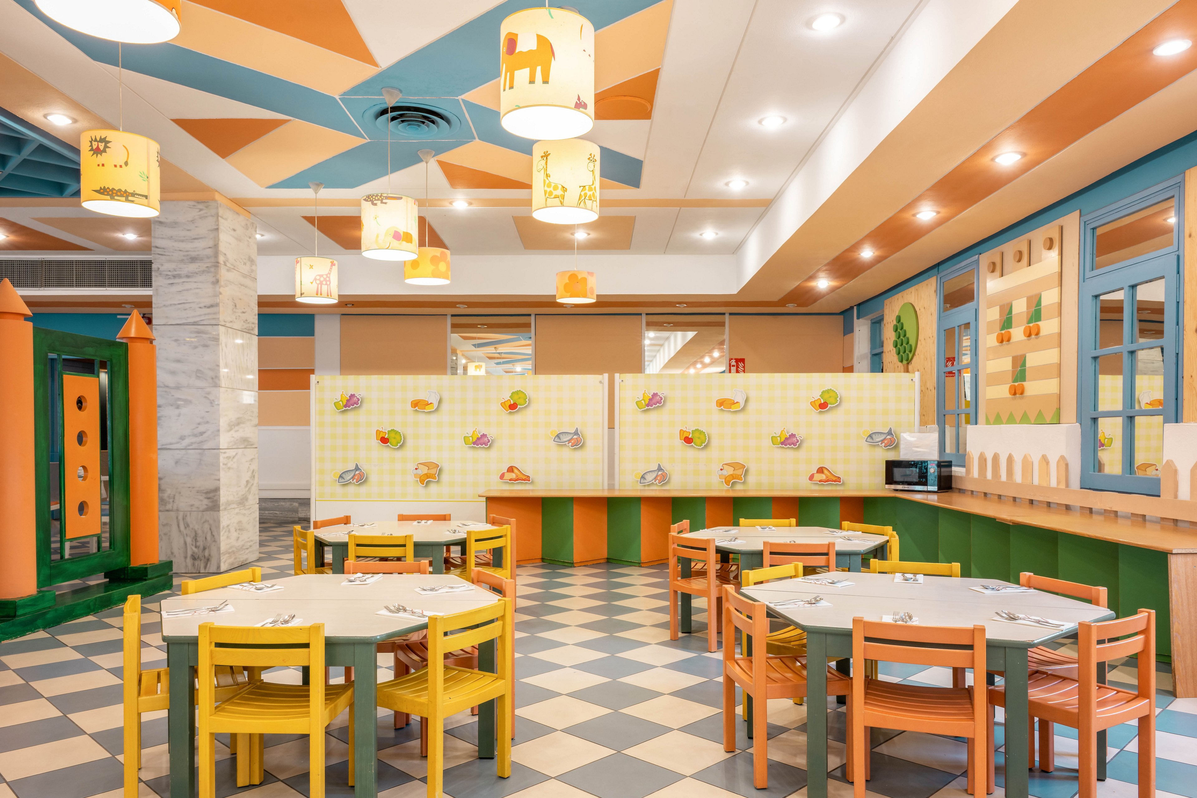 The image shows a brightly colored cafeteria designed for children, featuring small tables and chairs in orange, yellow, and green hues. The setting includes whimsical wall decorations and ceiling lights with animal illustrations, creating a cheerful and inviting atmosphere. The geometric floor tiles and pastel palette enhance the playful mood, while the space appears clean and well-organized for group activities or meals.