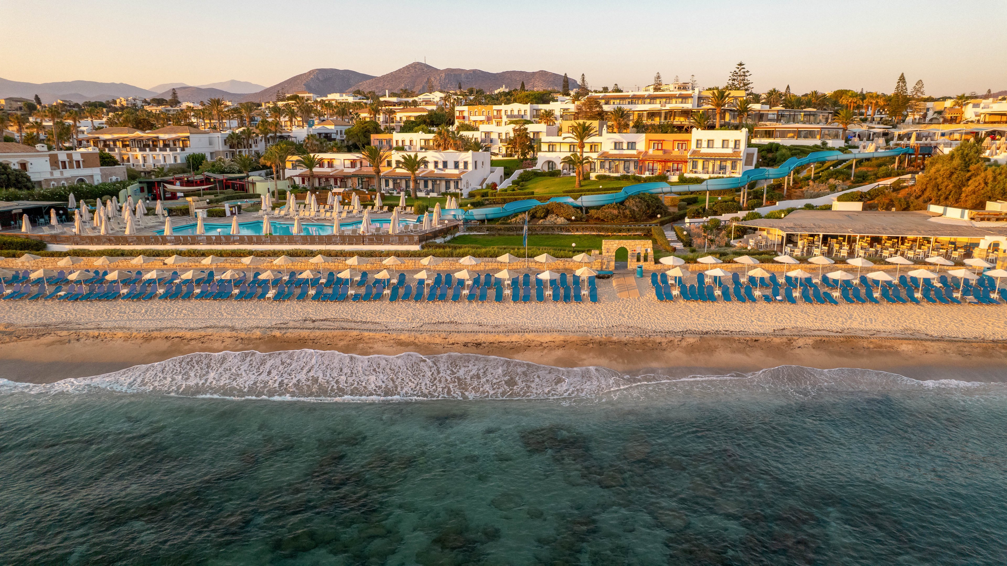 This image shows a luxury beachfront resort captured from above at sunset. Rows of blue sun loungers and umbrellas line the sandy shore, with a large swimming pool and landscaped gardens visible behind them. The resort buildings are surrounded by palm trees and greenery, with mountains in the background. The calm sea and clear sky create a tranquil atmosphere.