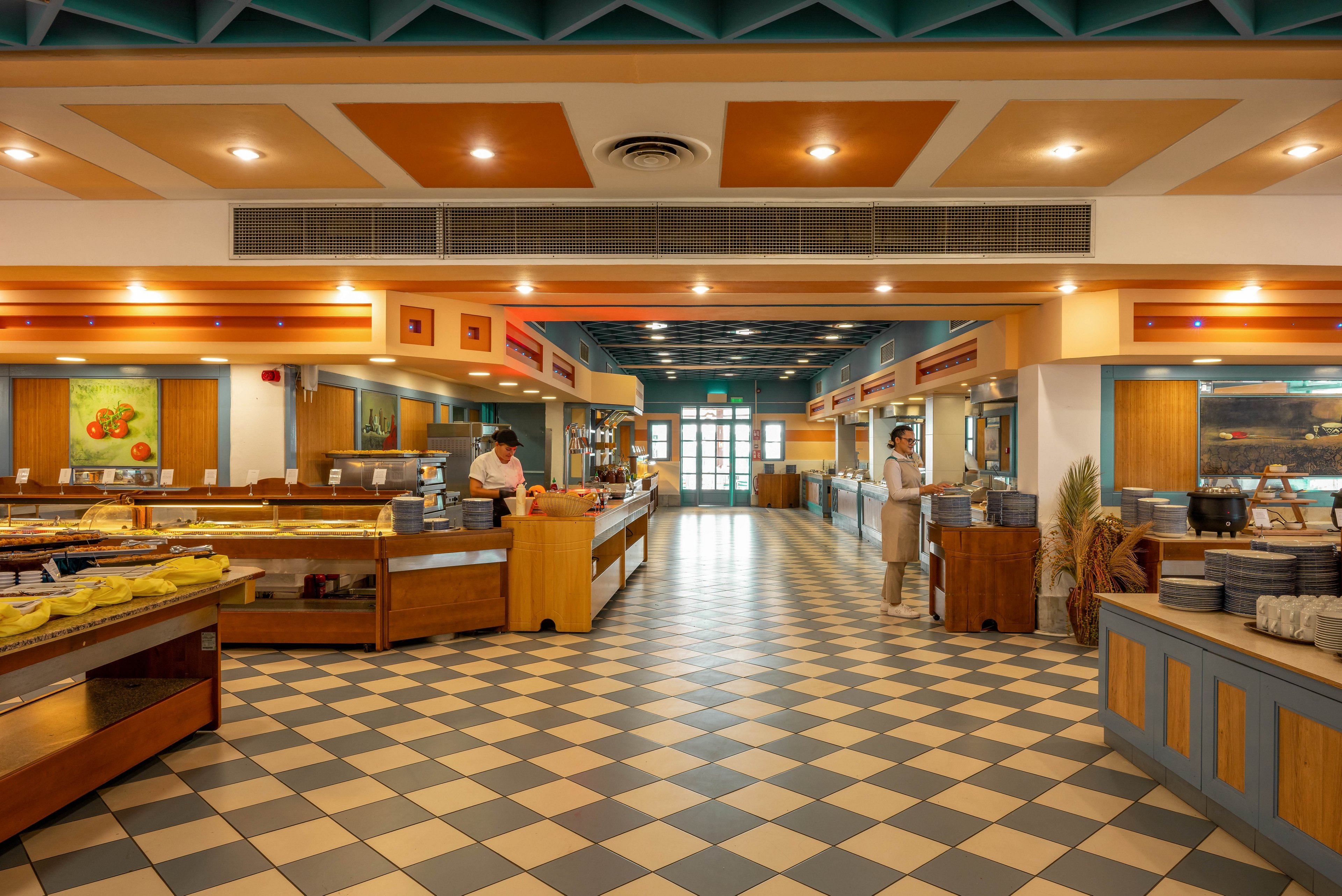 A wide-angle view of a brightly lit buffet restaurant features checkered tile flooring and wooden counters. Two staff members, dressed in uniforms, stand behind the serving stations, ready to assist guests. The interior is decorated with warm tones, geometric ceiling accents, and framed artwork on the walls. The setting appears clean and organized, with plates and food items arranged neatly. The overall mood is inviting and professional, suitable for family dining or group gatherings.